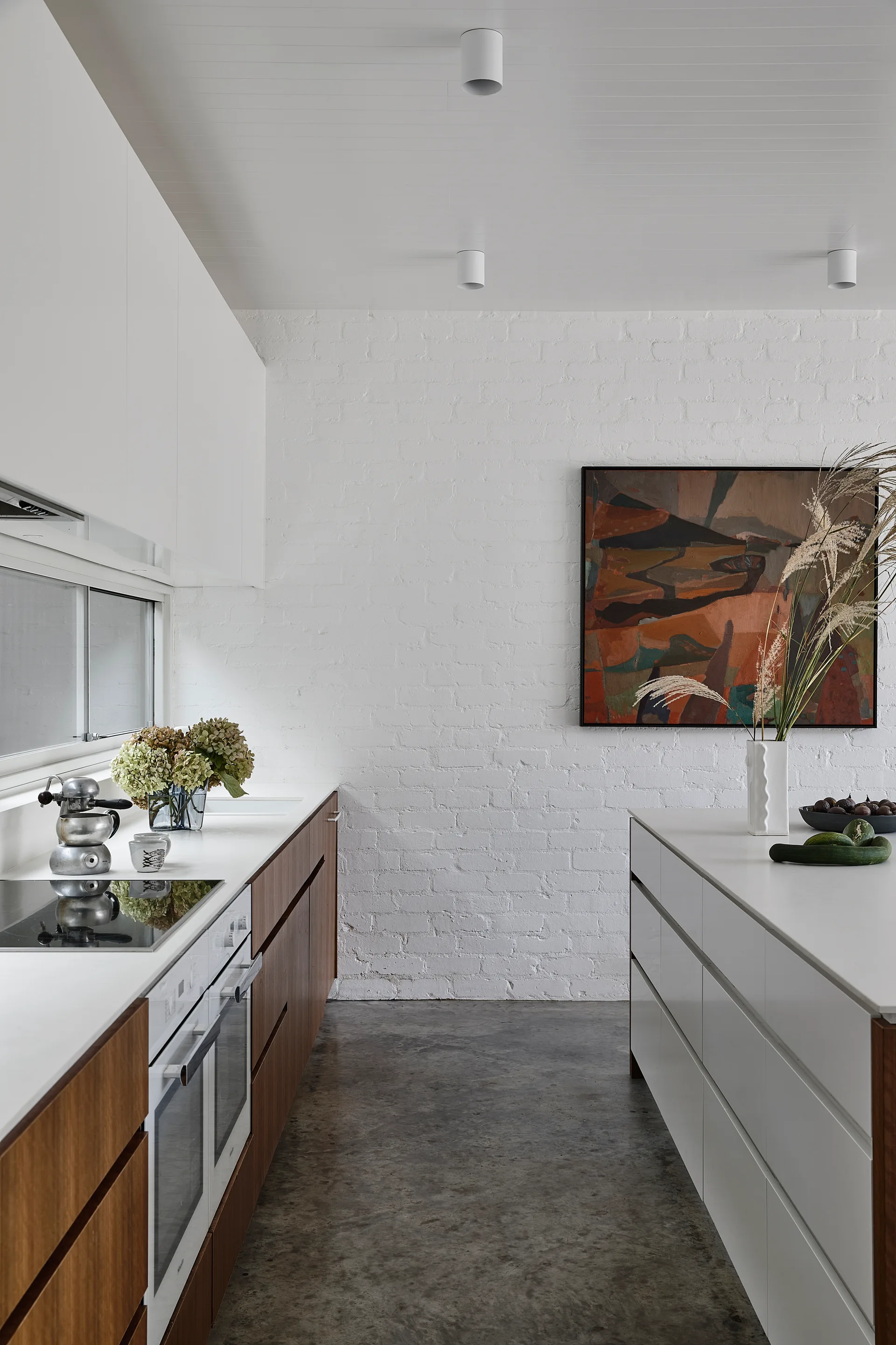 A modern kitchen features wood and white cabinetry, a polished concrete floor, and an abstract painting hung on a white painted brick wall.
