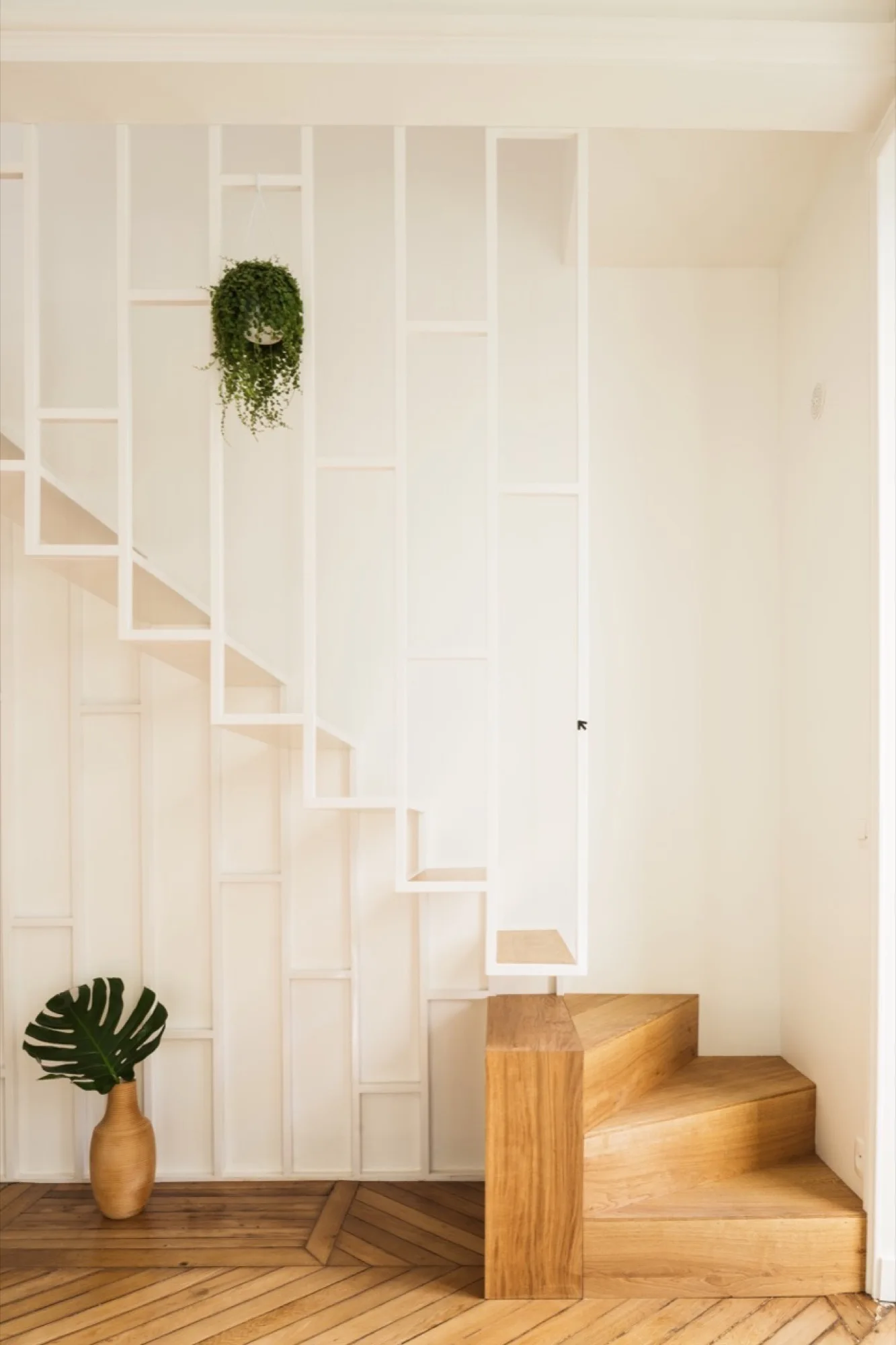 A minimalist staircase features light wood steps and a white geometric metal screen railing set against a clean white wall.