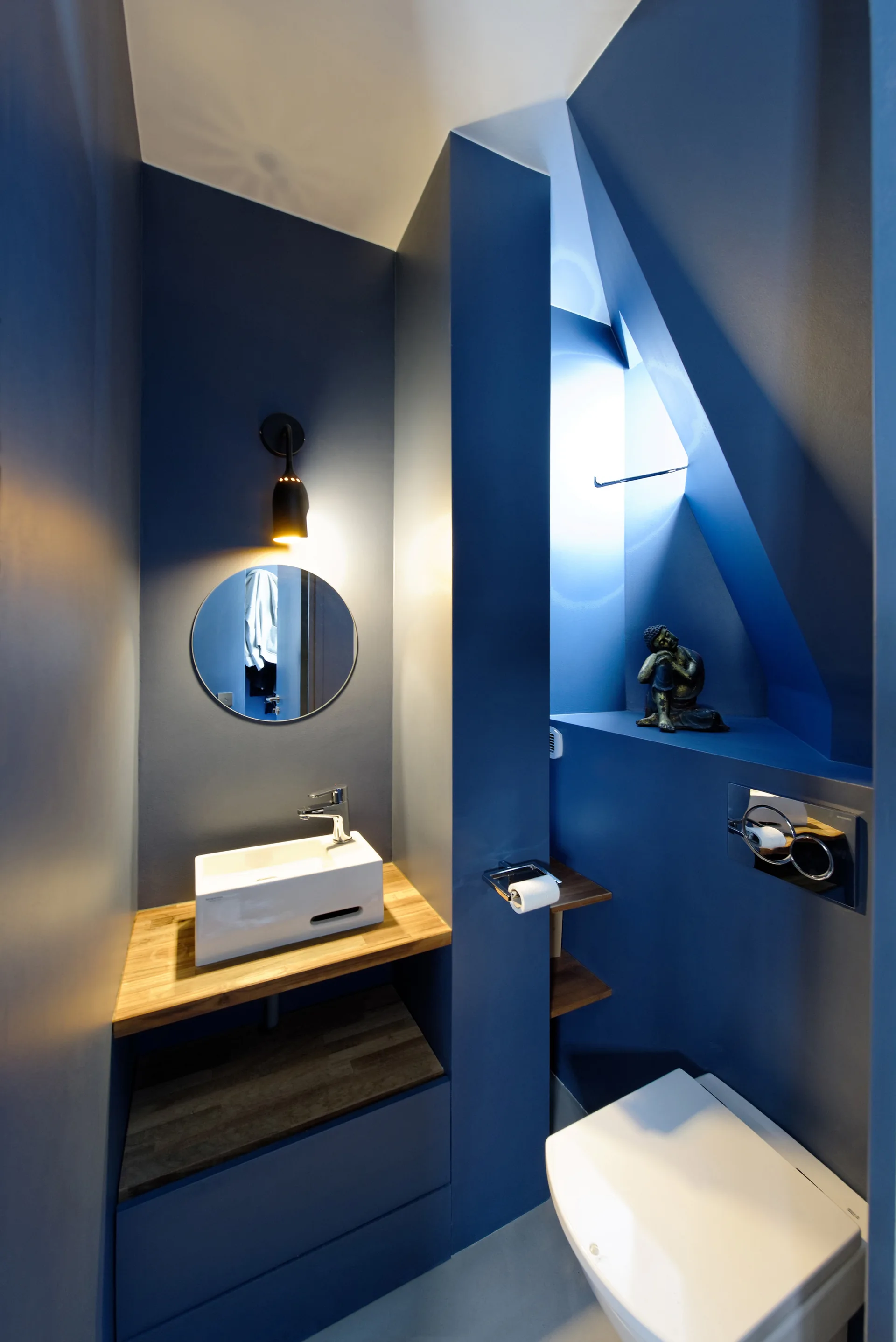 A small powder room features matte blue walls, a light wood floating vanity with a vessel sink, a round mirror, and a wall-mounted toilet.