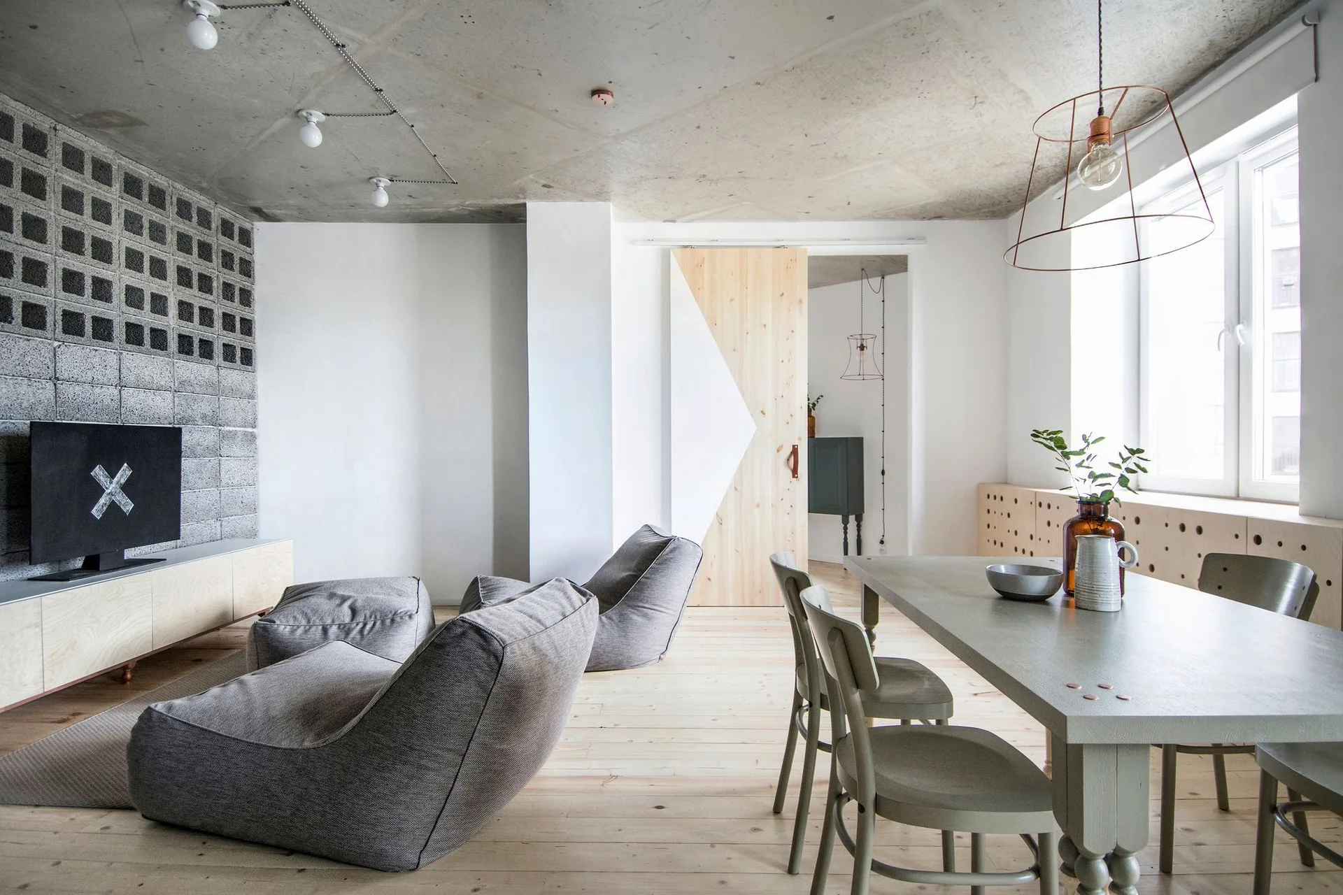 An open-plan living and dining room featuring an exposed concrete ceiling, light wood floors, grey beanbag chairs, and a textured cinder block wall.