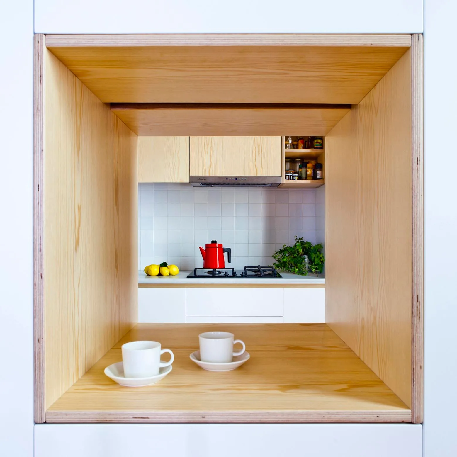 A view through a deep light wood frame onto a kitchen counter with a red kettle, white tile backsplash, and light wood upper cabinets.
