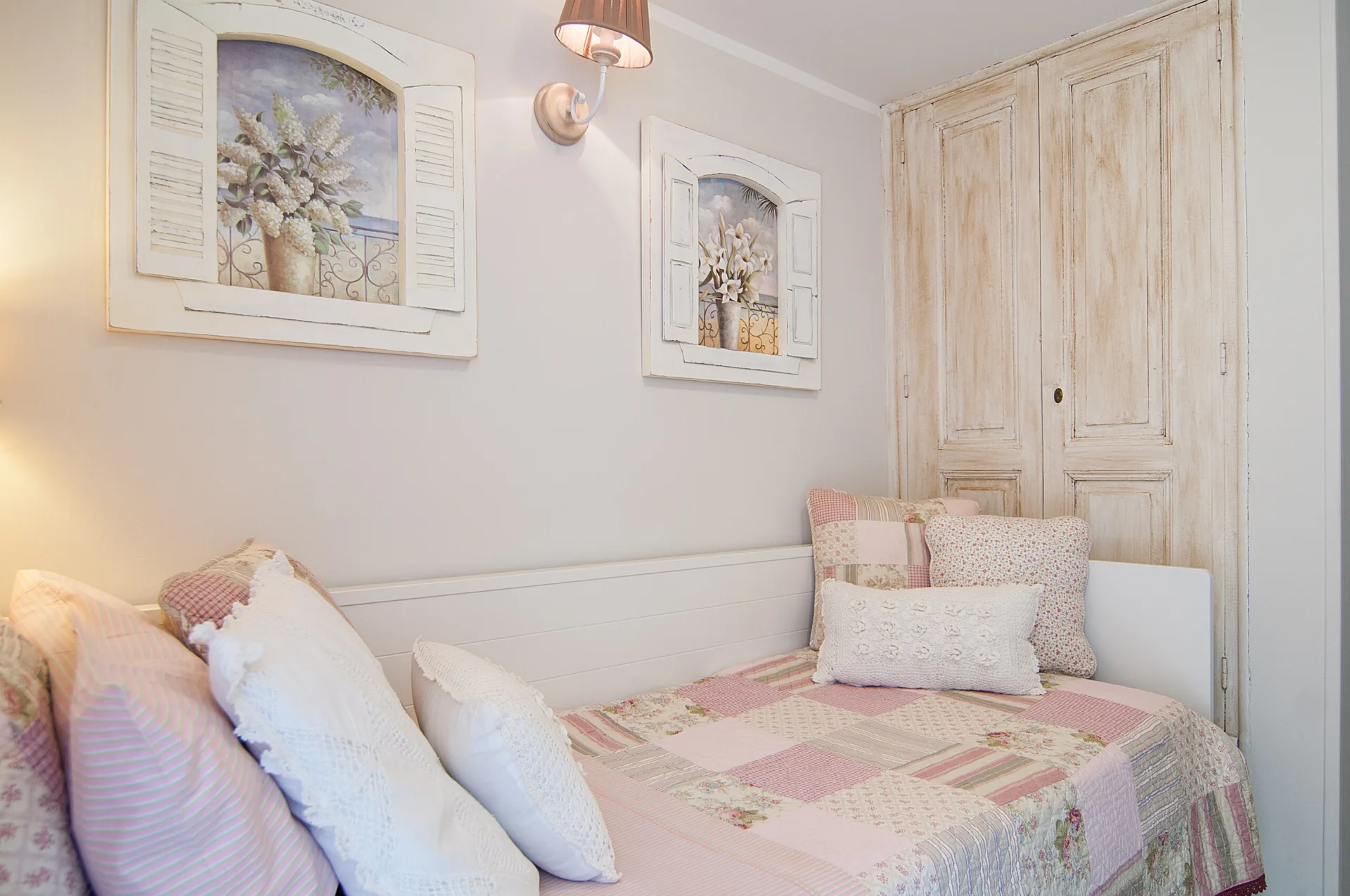 A cottage-style daybed with pink patchwork bedding and pillows is nestled next to a distressed wood wardrobe and framed shutter wall art.
