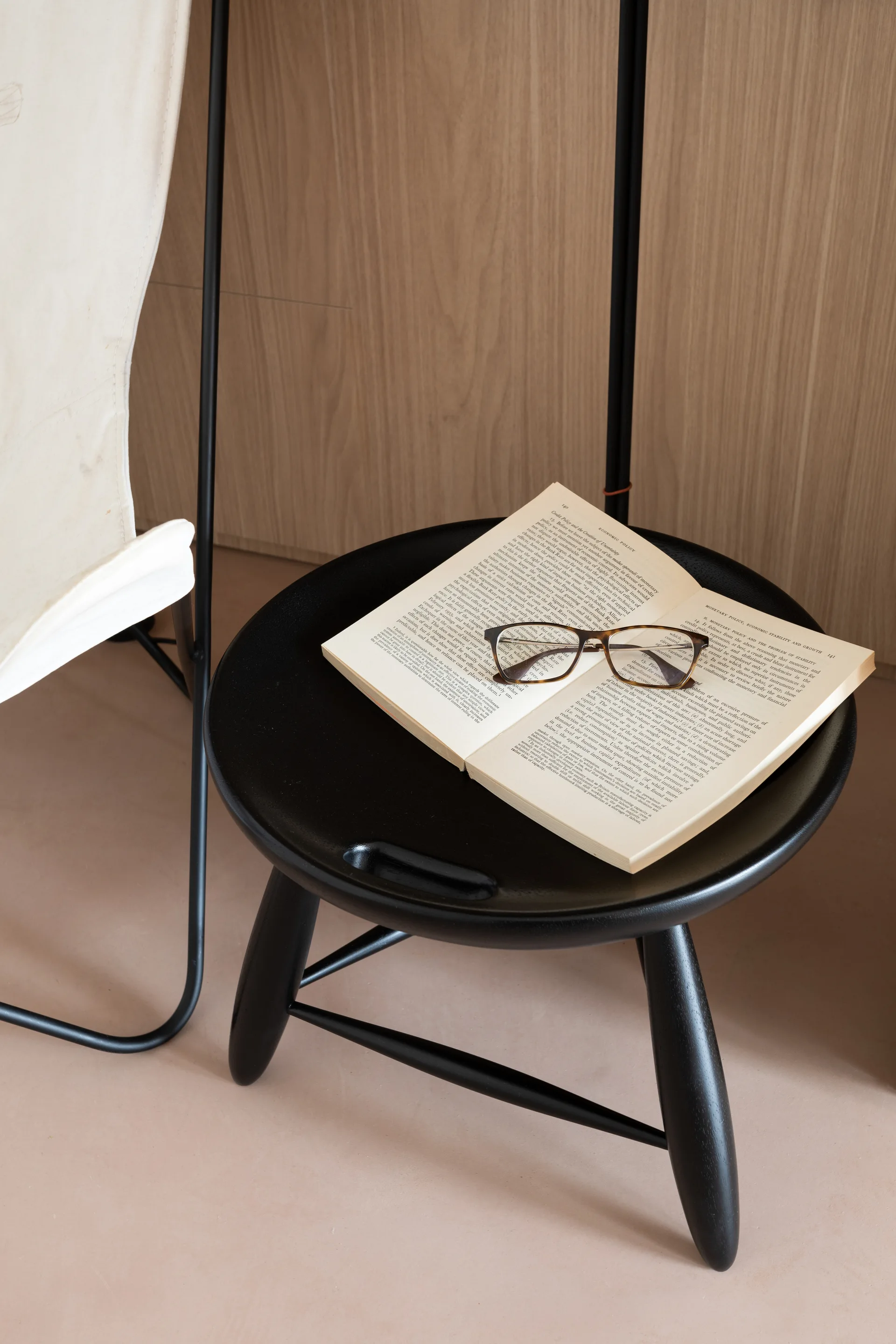 A black wooden stool with a recessed handle holds an open book and glasses, positioned next to a canvas chair against vertical wood paneling.