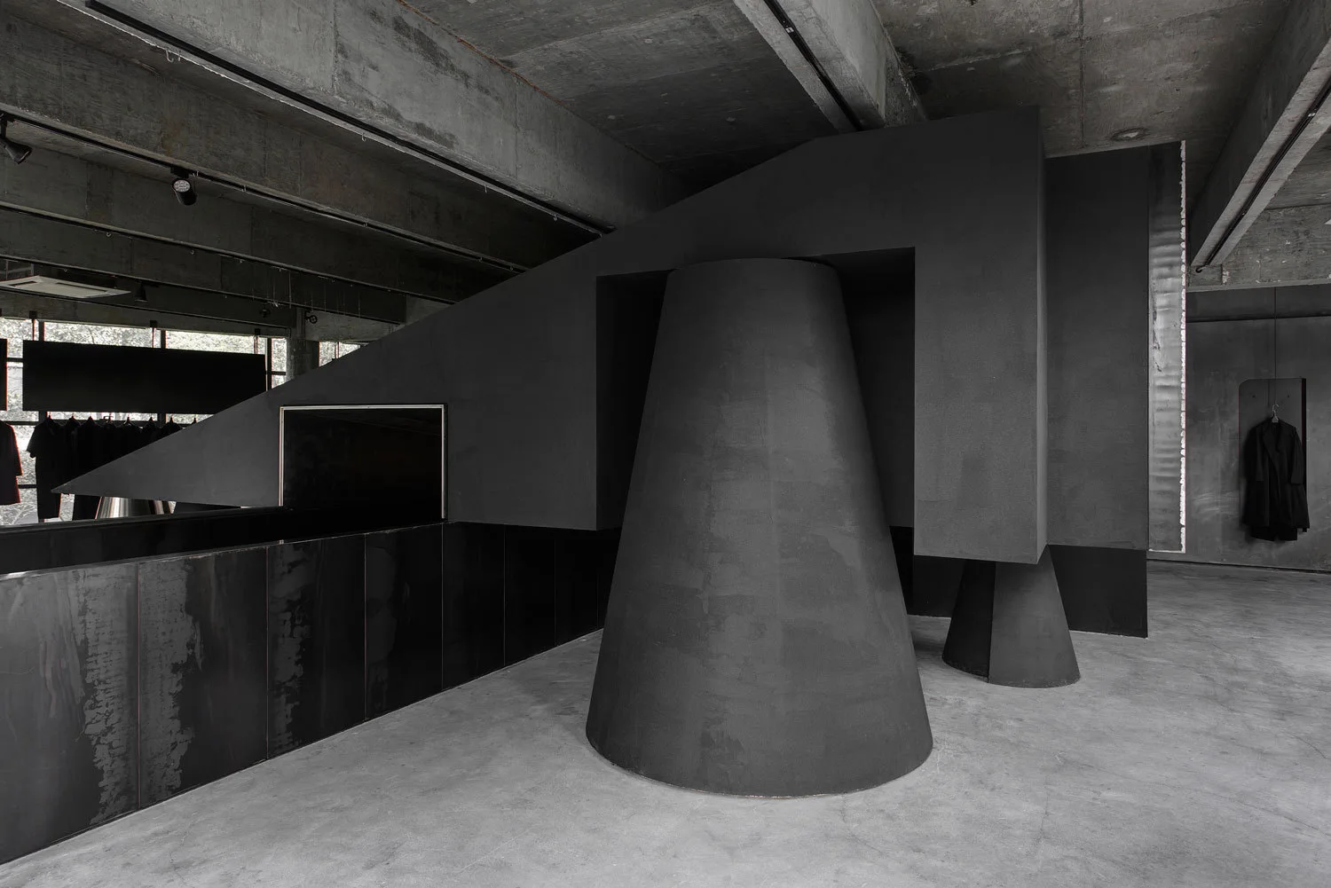 A minimalist brutalist interior featuring massive matte black conical and angular sculptural installations against raw exposed concrete ceiling and floor.