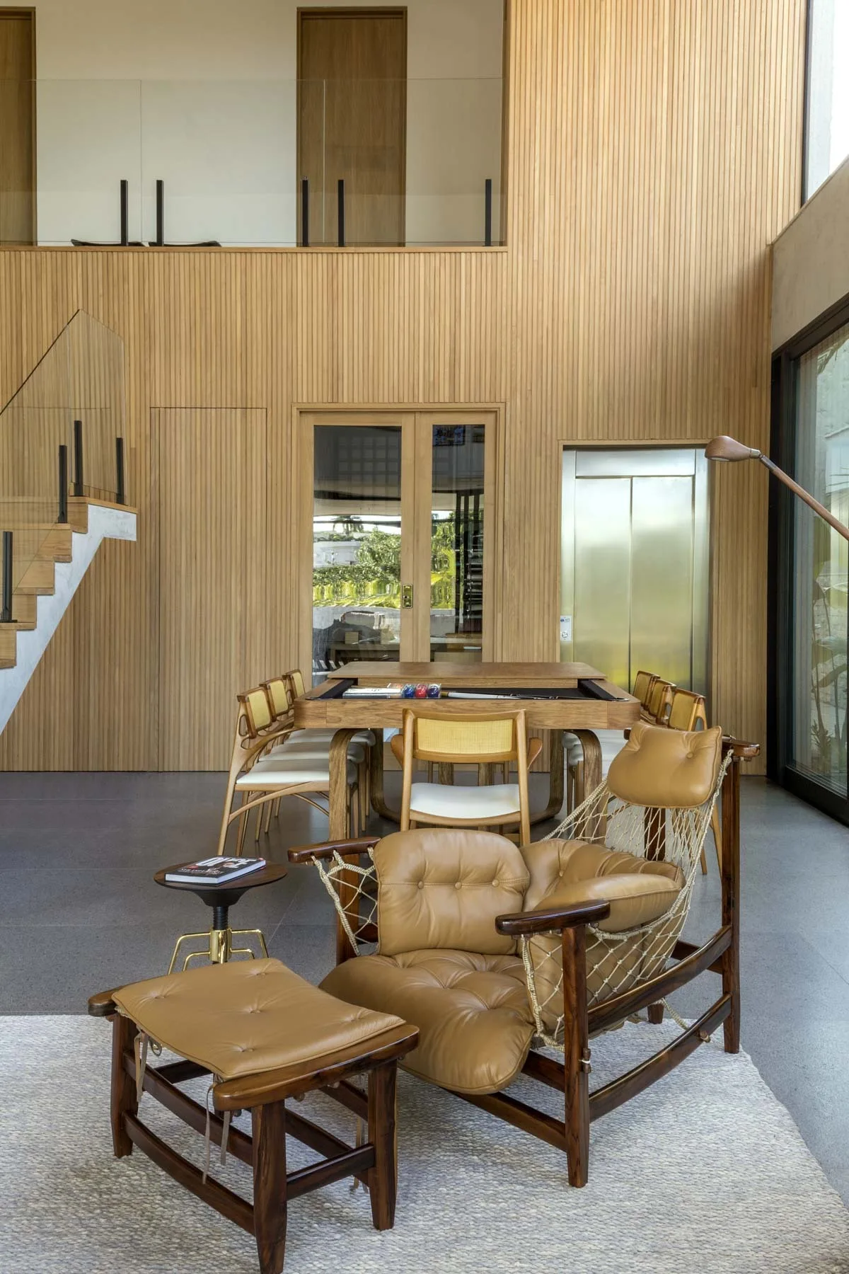 A double-height dining room features vertical wood-slat walls, a wooden dining table with chairs, and a tufted leather lounge chair on a textured rug.