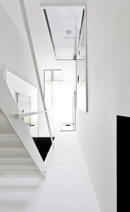 A minimalist white staircase features a simple metal handrail, sharp geometric angles, and rectangular wall mirrors reflecting light from a skylight.
