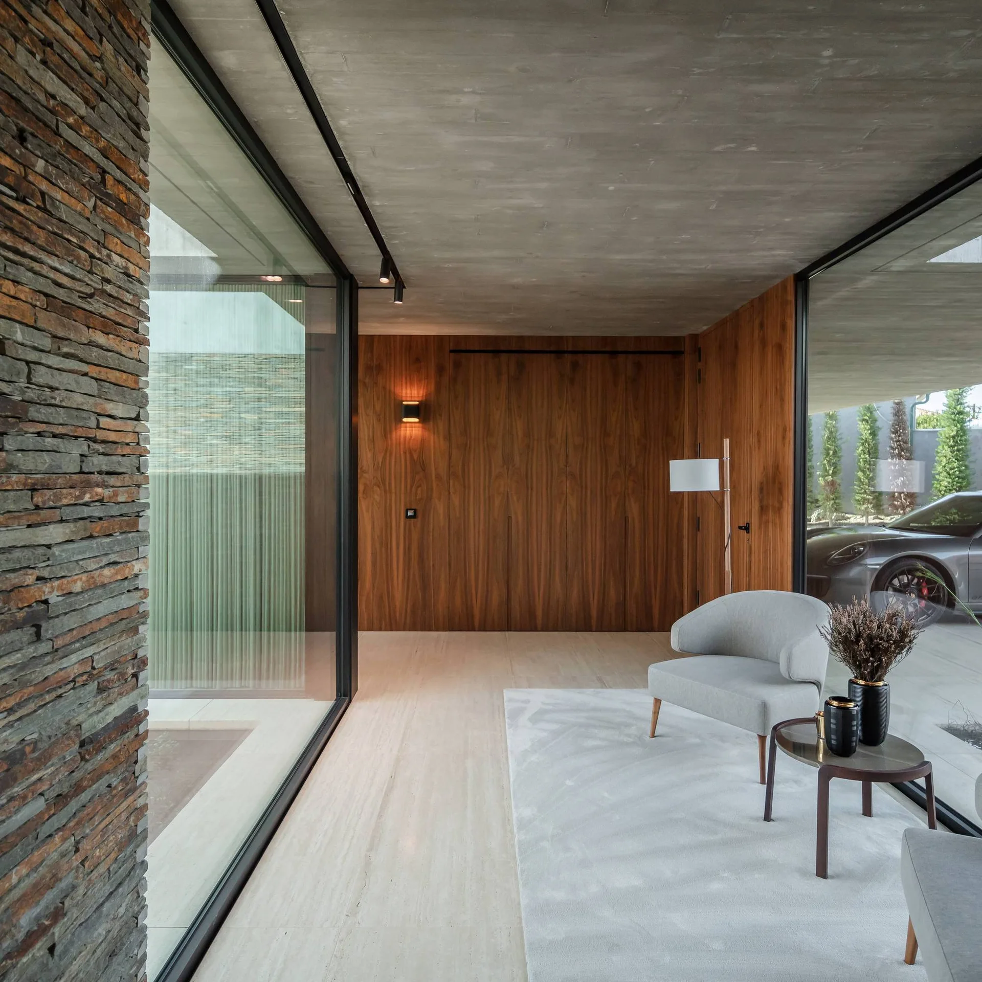 A minimalist lounge features a grey armchair on a light rug, surrounded by large glass windows, a wood-paneled wall, and a raw concrete ceiling.