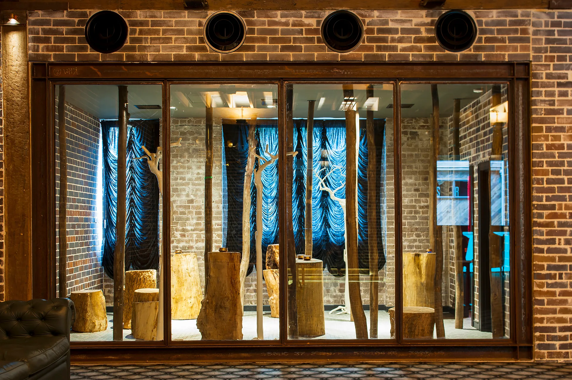 A steel-framed glass partition wall showcases natural wood stump stools and decorative branches against draped blue curtains and exposed brick.