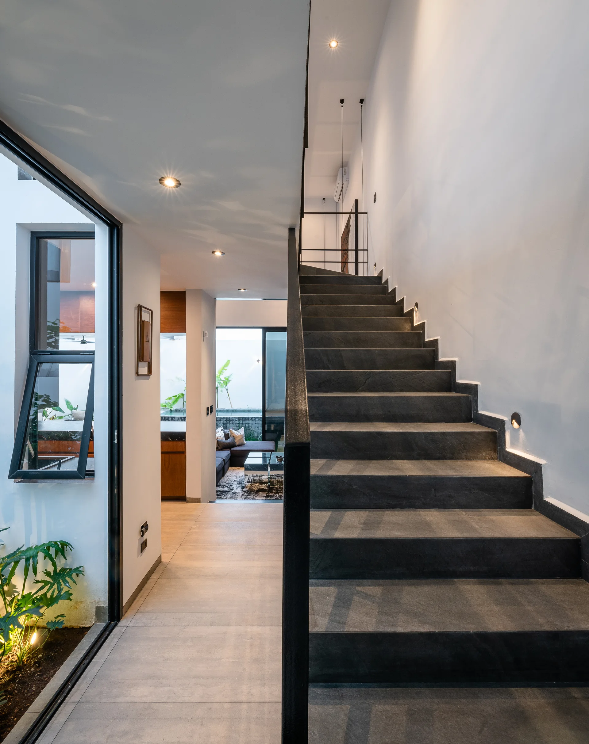 A modern hallway features a dark grey stone staircase with minimal black handrails next to an indoor lightwell window showing green plants.
