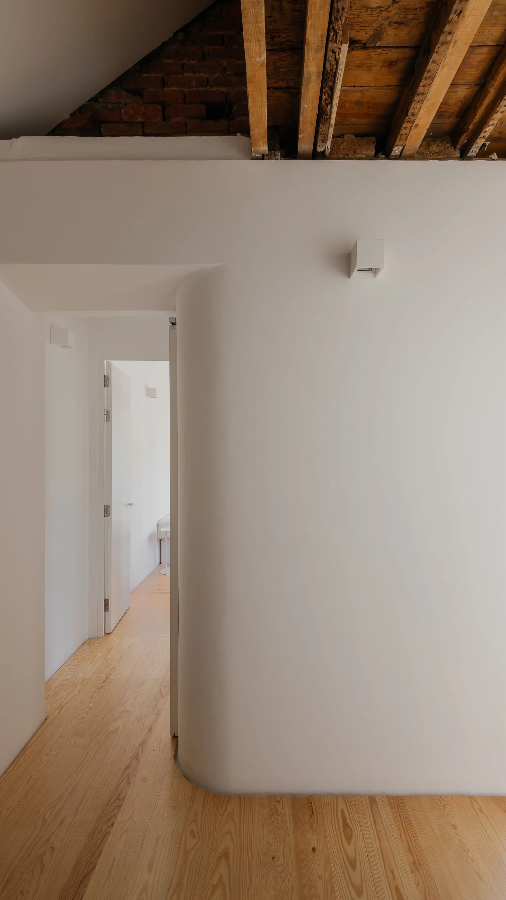 A minimalist white hallway features a prominent curved wall, light wood flooring, and an unfinished ceiling with exposed timber beams and brick.