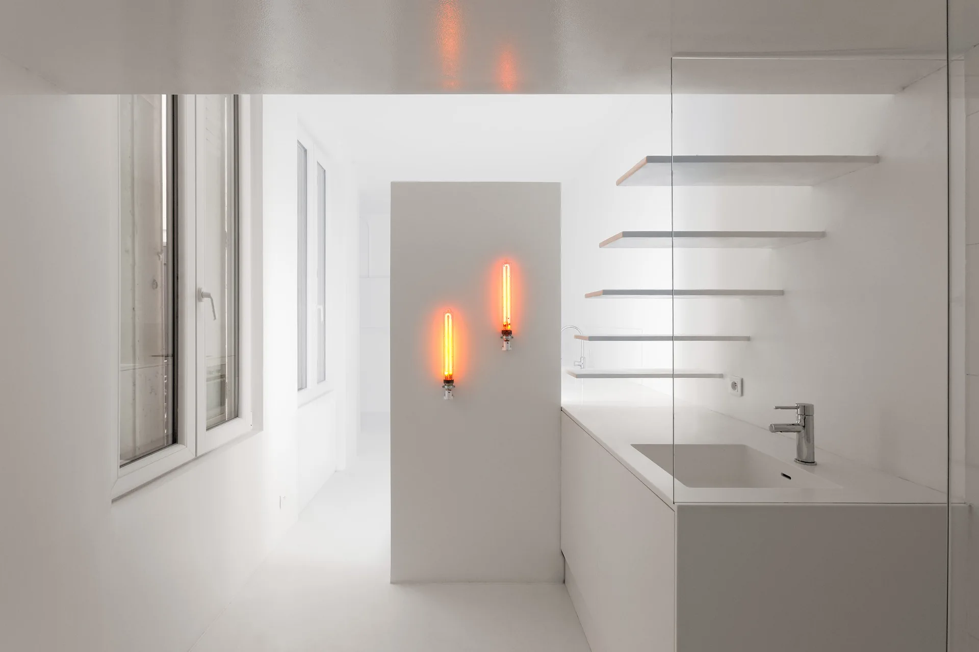 A minimalist white bathroom features a built-in vanity, a glass partition, floating shelves, and two glowing orange vertical tube lights.