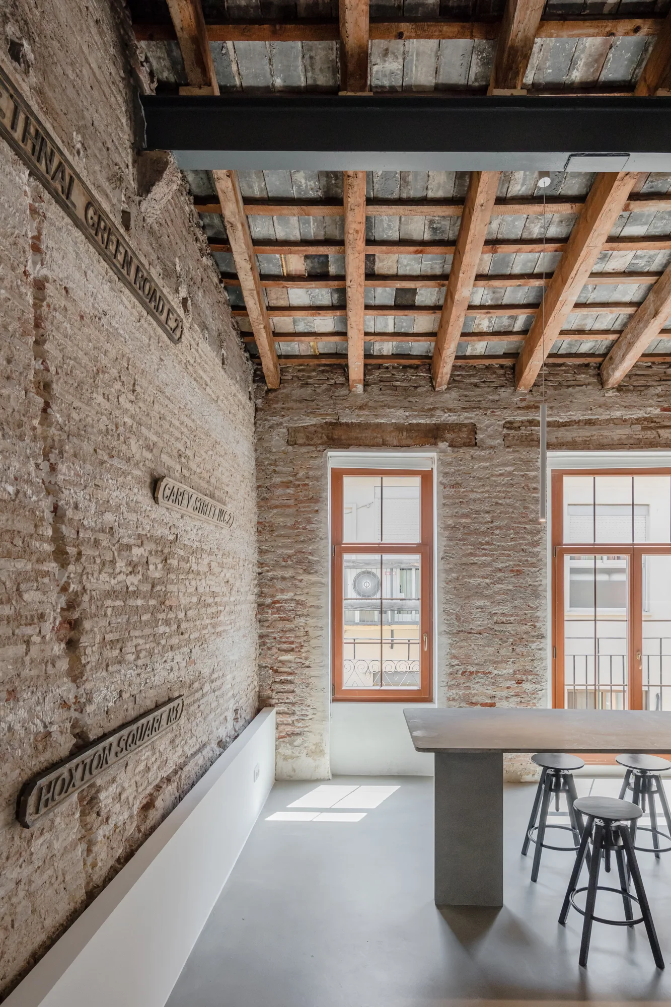 An industrial dining room features a stone table with metal stools, exposed brick walls with vintage street signs, and a wood-beamed ceiling.