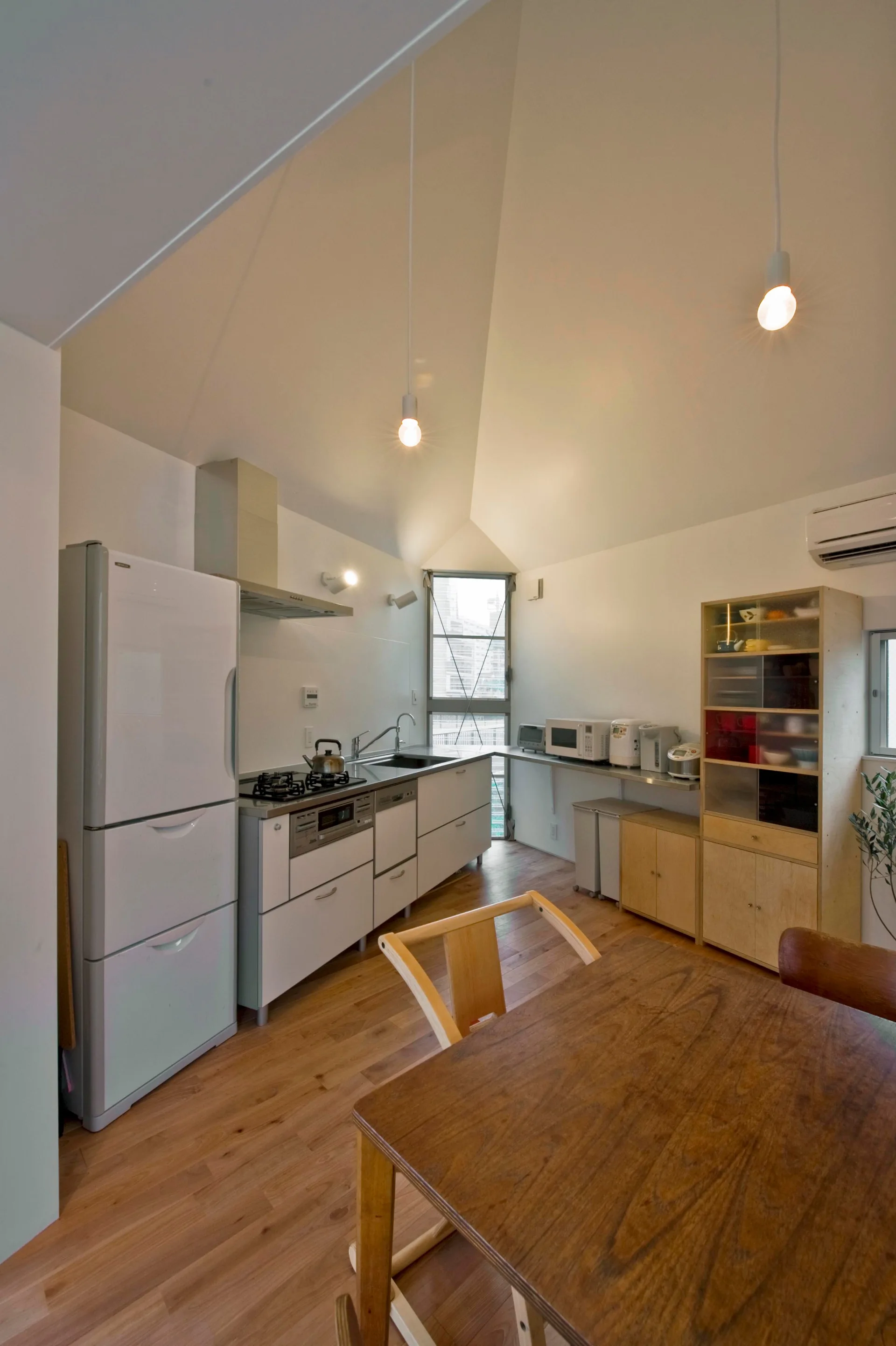 A minimalist kitchen features a vaulted ceiling, white cabinetry, a freestanding refrigerator, a wooden dining table, and a narrow vertical window.