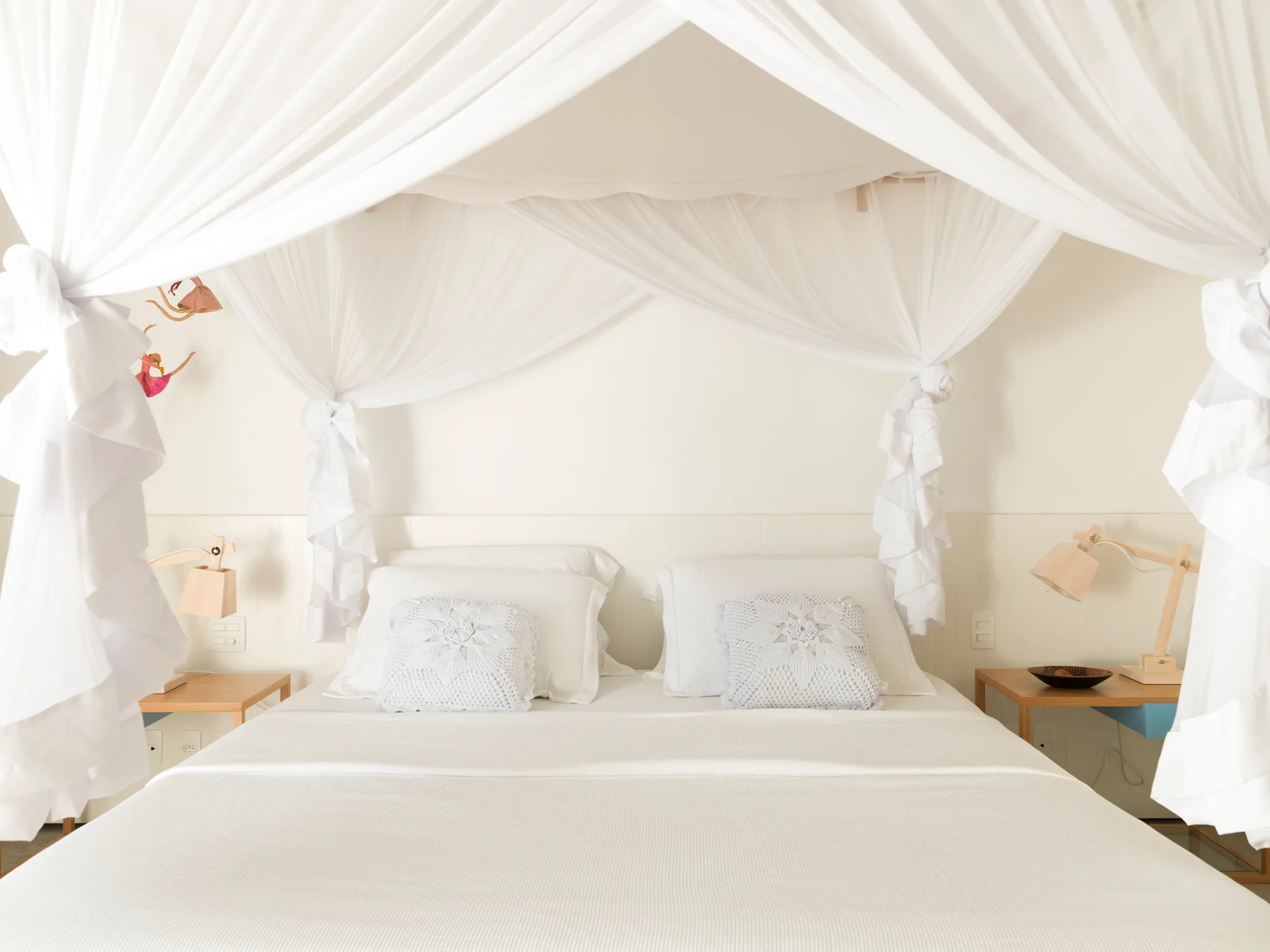 A white canopy bed features draped sheer white curtains, white bedding with two crochet throw pillows, and light wood nightstands on each side.