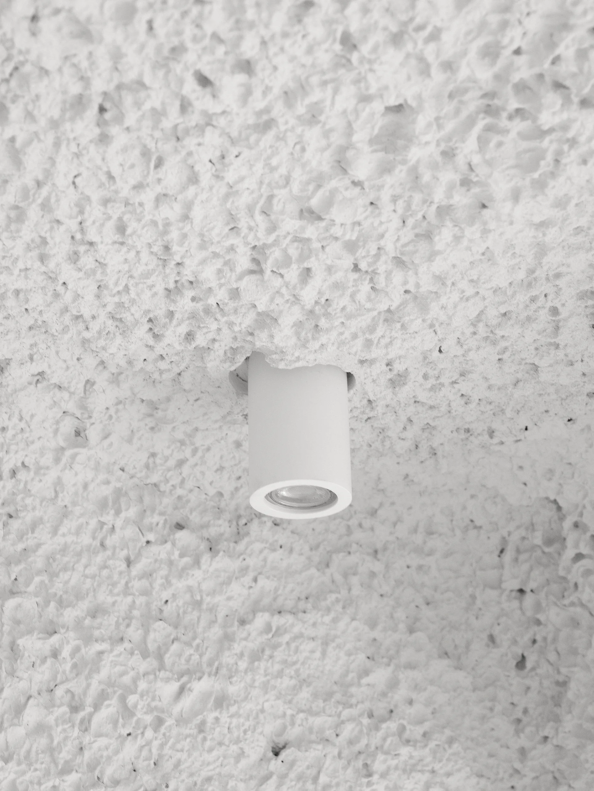 A close-up of a matte white cylindrical spotlight recessed into a highly textured, rough white plaster ceiling.