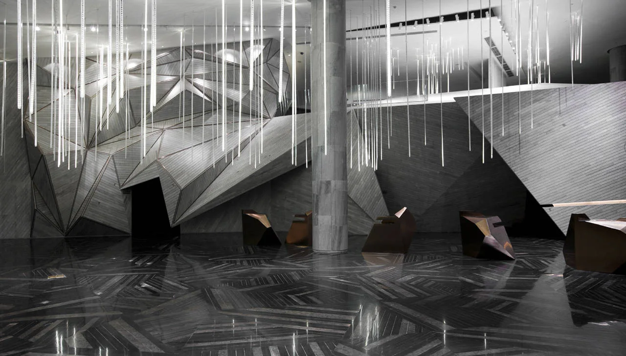 A spacious lobby featuring faceted geometric wood wall panels, vertical glass pendant lights, and a highly polished dark patterned stone floor.