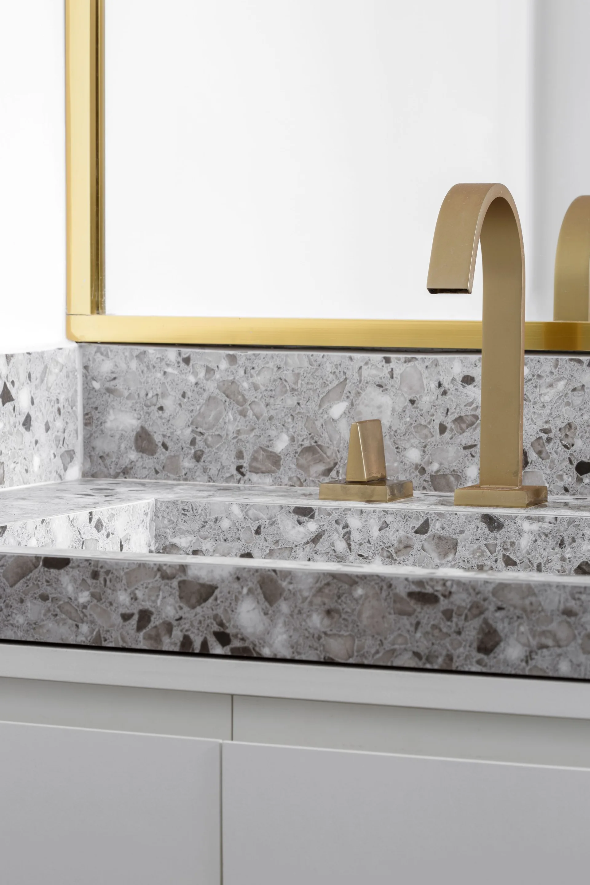 A close-up of a bathroom vanity featuring an integrated grey terrazzo sink, matching backsplash, satin gold faucet, and a gold-framed mirror.