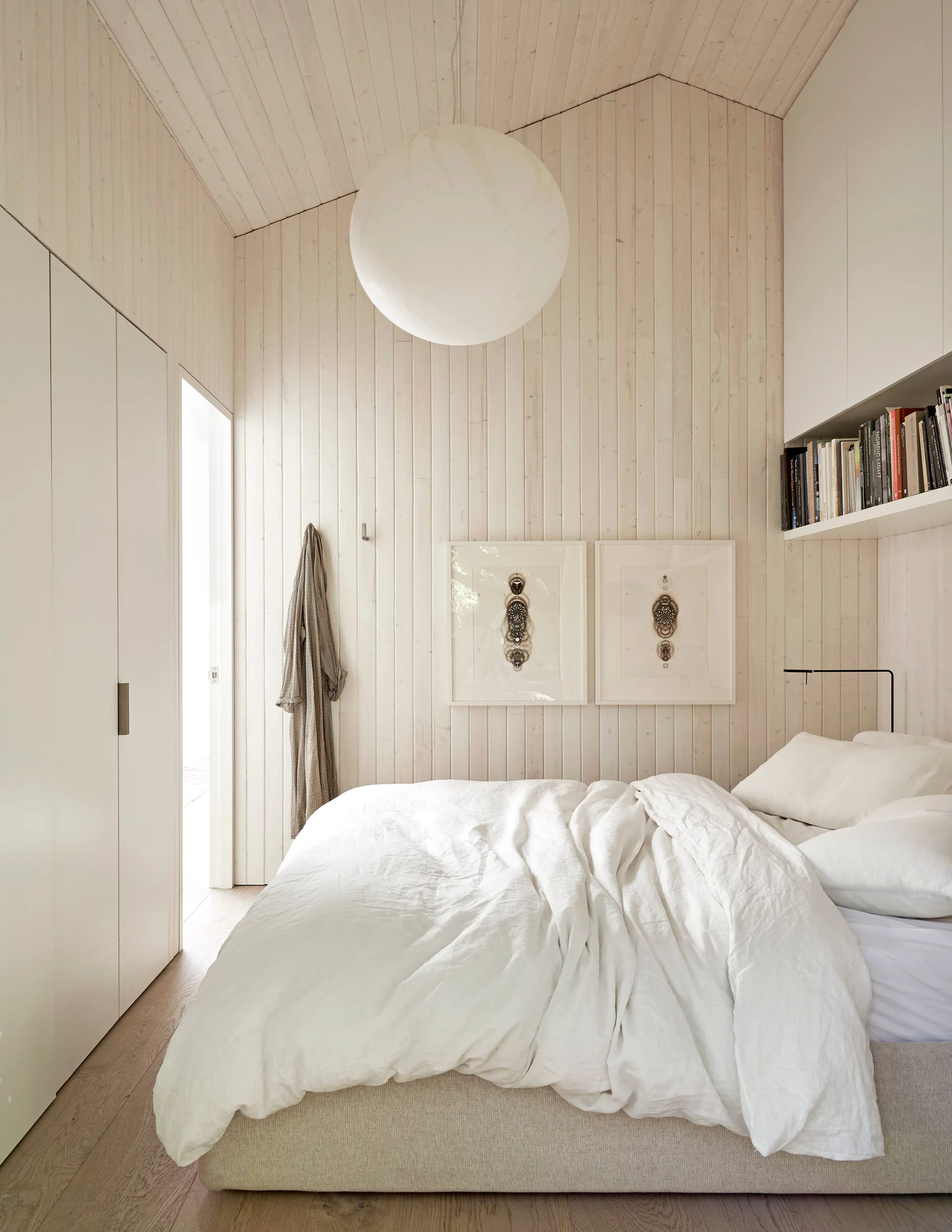 A minimalist bedroom features whitewashed vertical wood paneled walls, a vaulted ceiling, a bed with white linen sheets, and a large round pendant light.