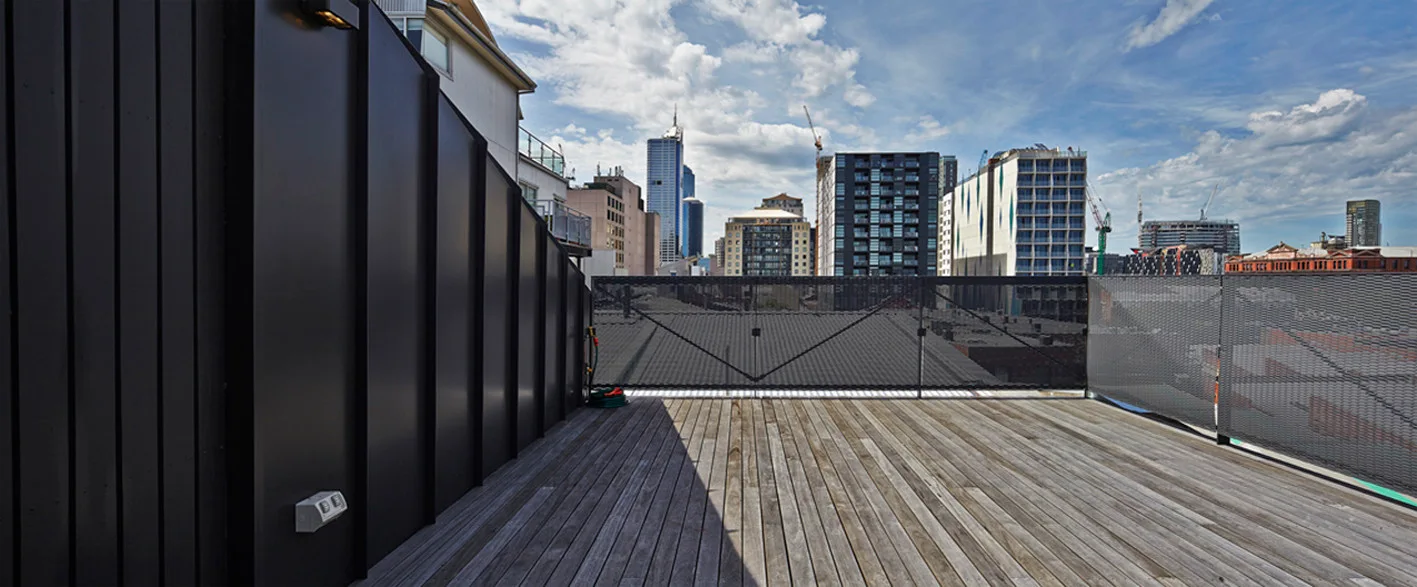 An outdoor rooftop terrace features weathered wooden plank flooring, a dark vertical metal privacy wall, and a mesh perimeter railing against a city skyline.