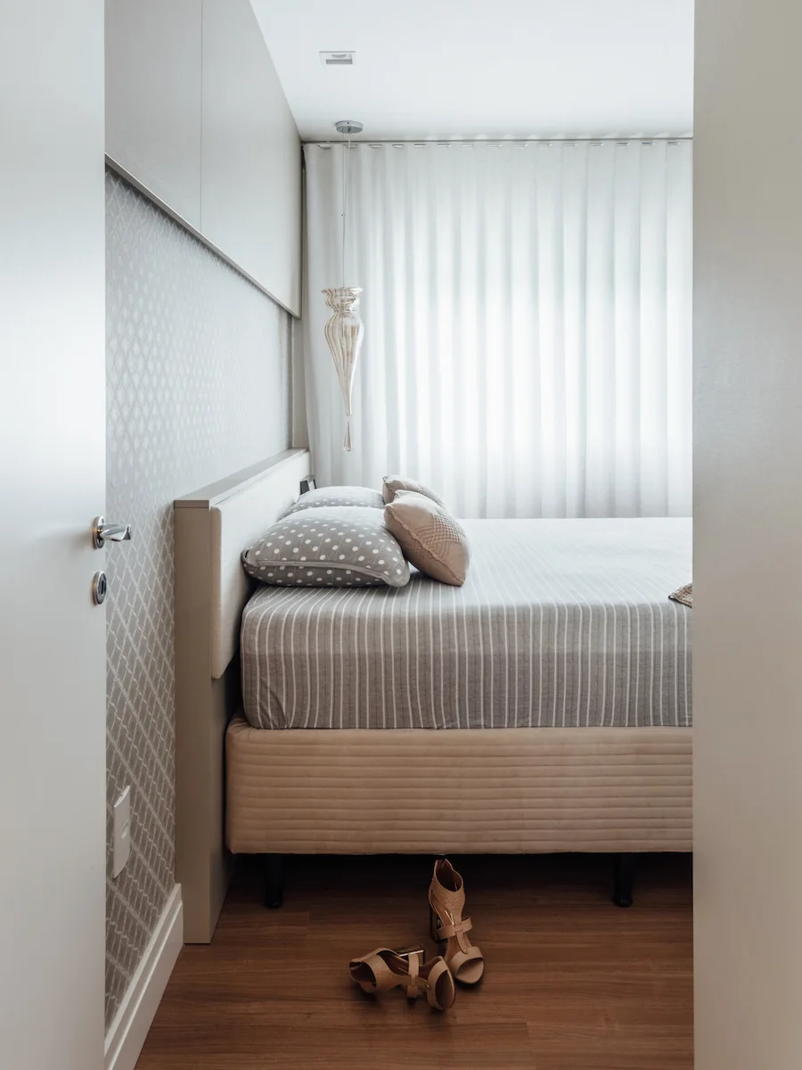 A compact bedroom features an upholstered bed with patterned pillows, overhead built-in cabinets, and a geometric wallpaper accent wall.