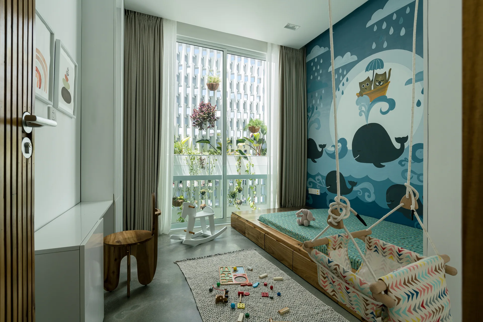 A children's bedroom features a large blue whale wall mural, a low wooden platform bed, a suspended fabric swing, and a concrete floor.