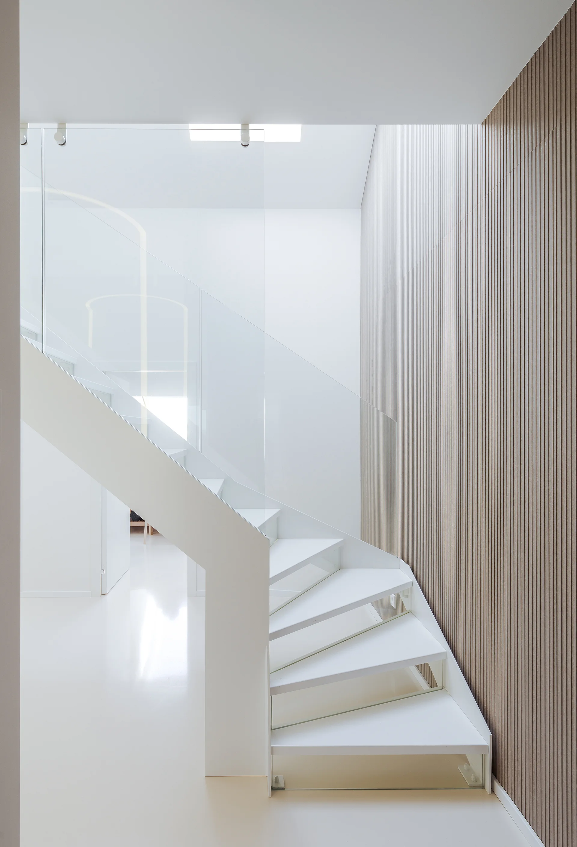 A modern white helical staircase with a clear glass railing ascends next to a wall clad in vertical wood-toned slats.
