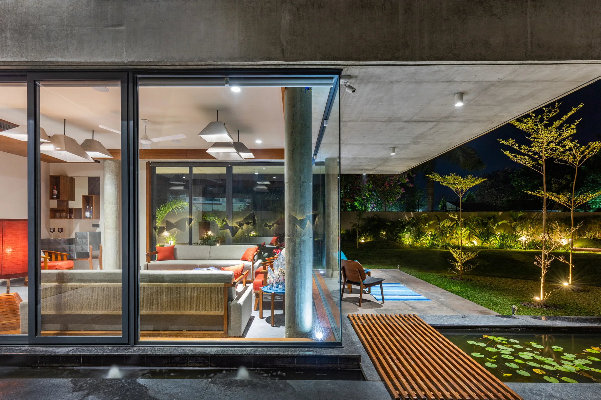 Floor-to-ceiling glass walls show a modern living room transitioning to a concrete patio, lawn, and small pond with lily pads.