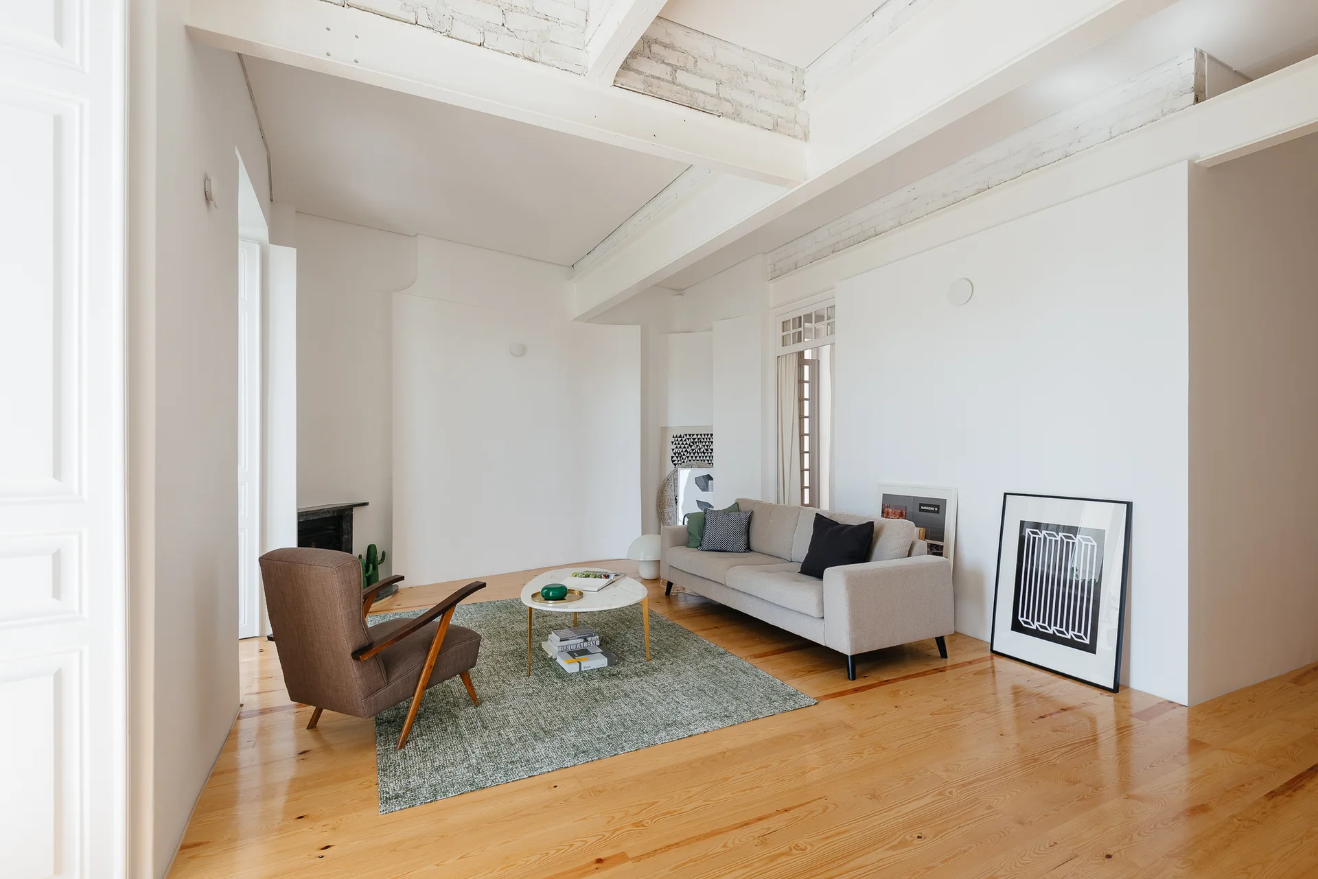 A minimalist living room with a grey sofa, brown armchair, and round coffee table on a green rug beneath white ceiling beams.