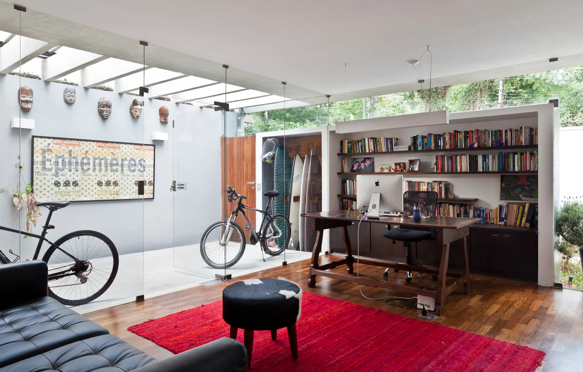 A modern home office features a wooden desk, a vibrant red area rug, and a floor-to-ceiling glass partition separating a gear storage area.