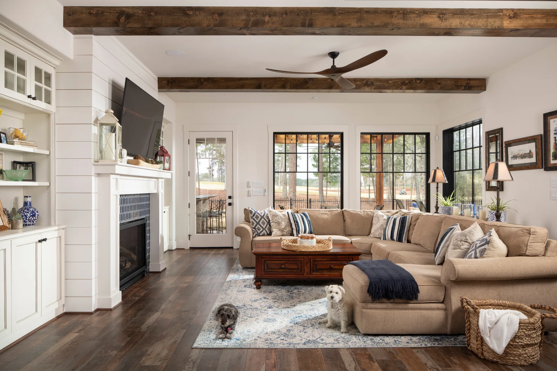A bright farmhouse living room featuring a beige sectional sofa, a fireplace with a white shiplap mantel, and dark exposed wood ceiling beams.