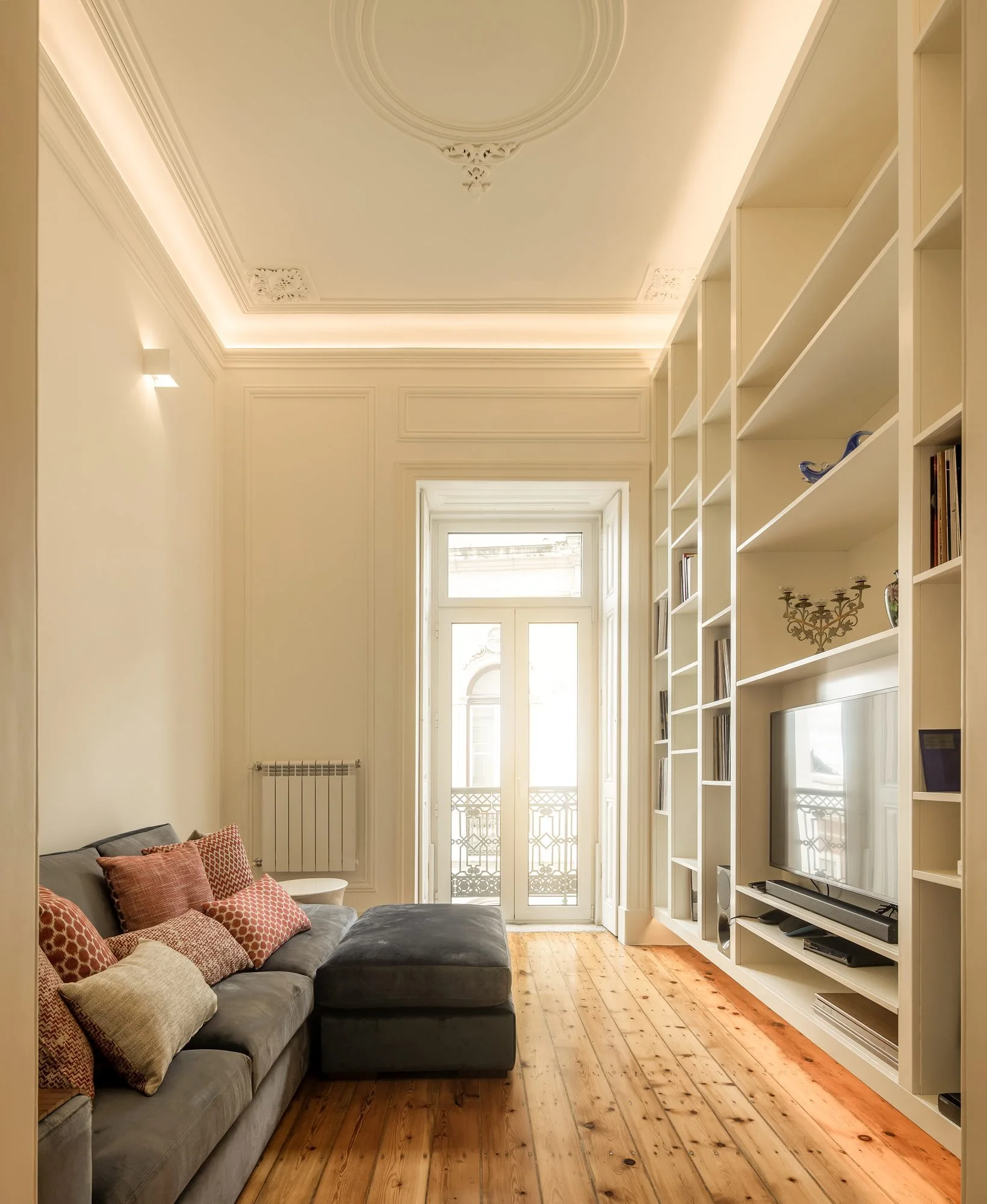 A narrow living room featuring a grey sectional sofa, custom white floor-to-ceiling bookshelves, and ornate ceiling molding with warm cove lighting.