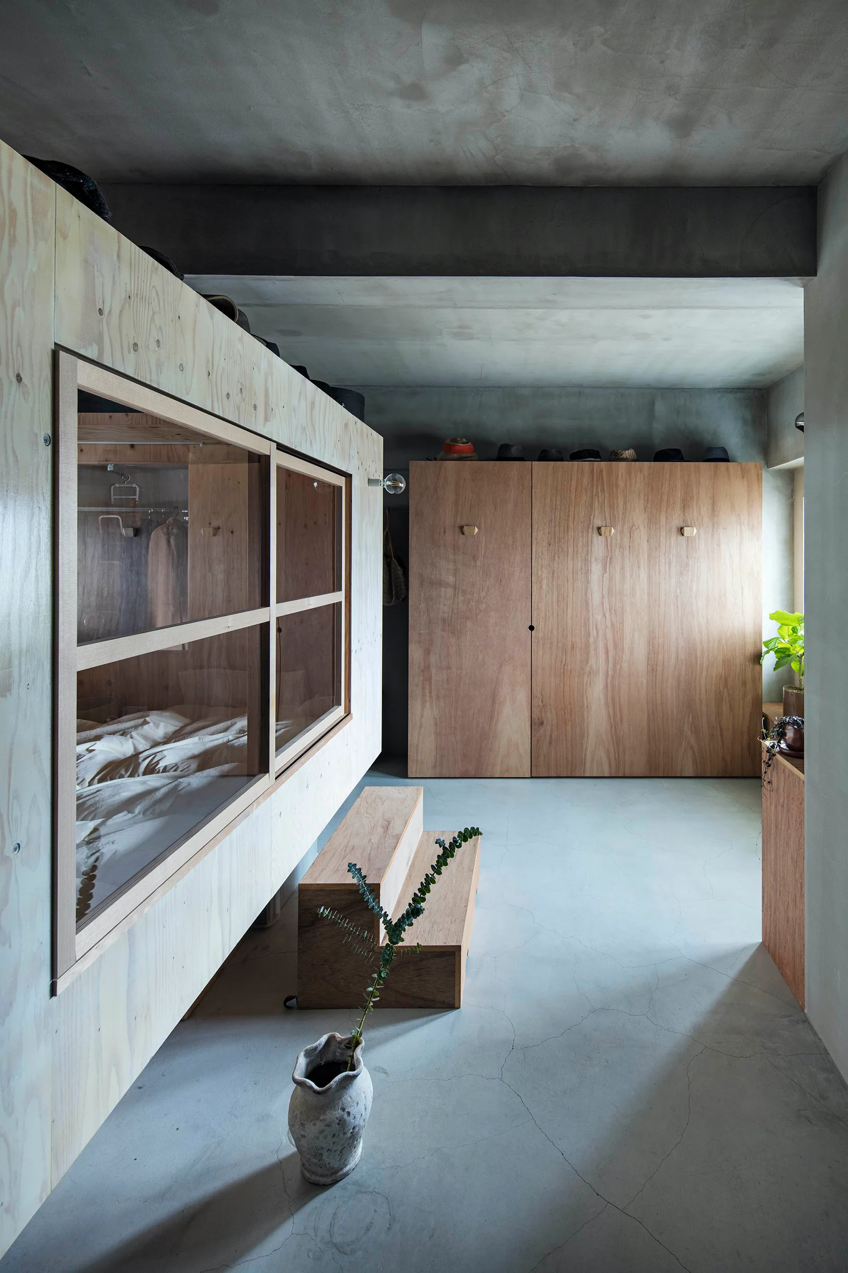 A minimalist bedroom featuring an elevated plywood sleeping cabin and a matching wardrobe set against raw concrete walls and floors.