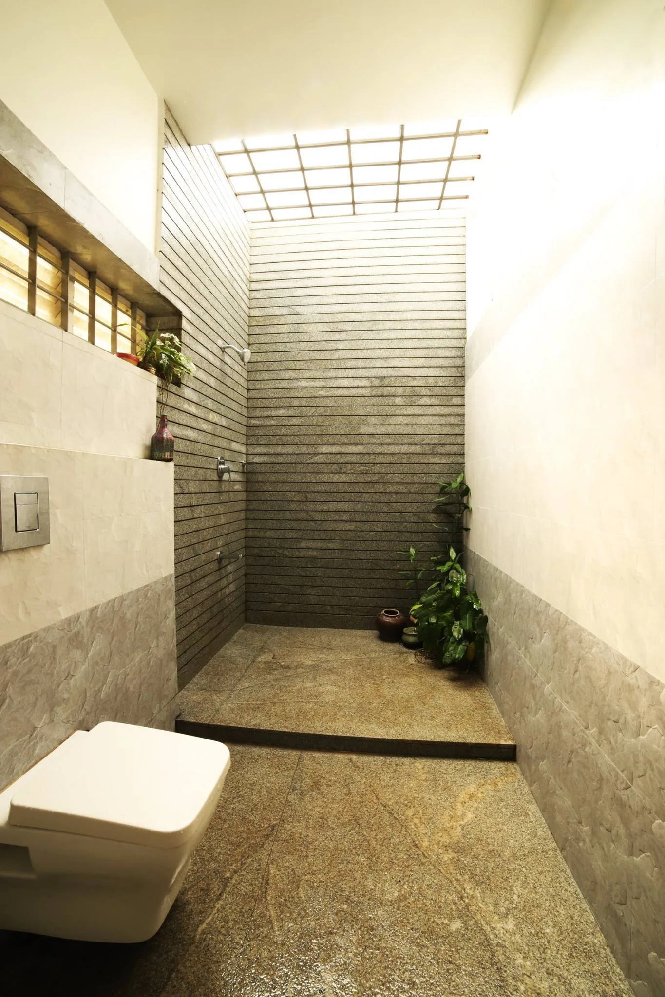 A modern bathroom features a wall-mounted toilet next to a walk-in shower with horizontal-slatted stone walls and a grid skylight.