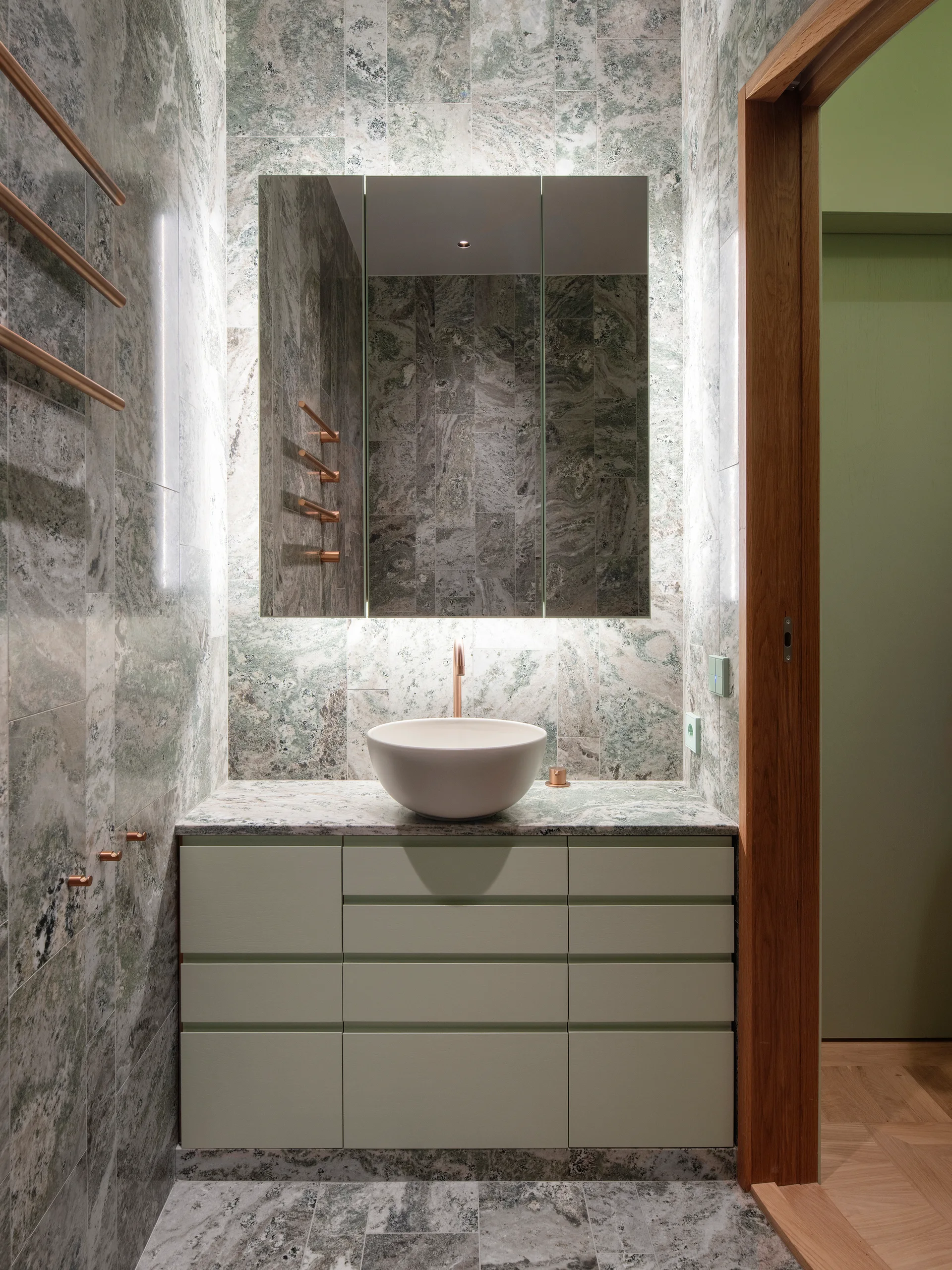A modern bathroom features green veined marble tiled walls, a round white vessel sink atop a sage green vanity, and minimalist copper metal accents.