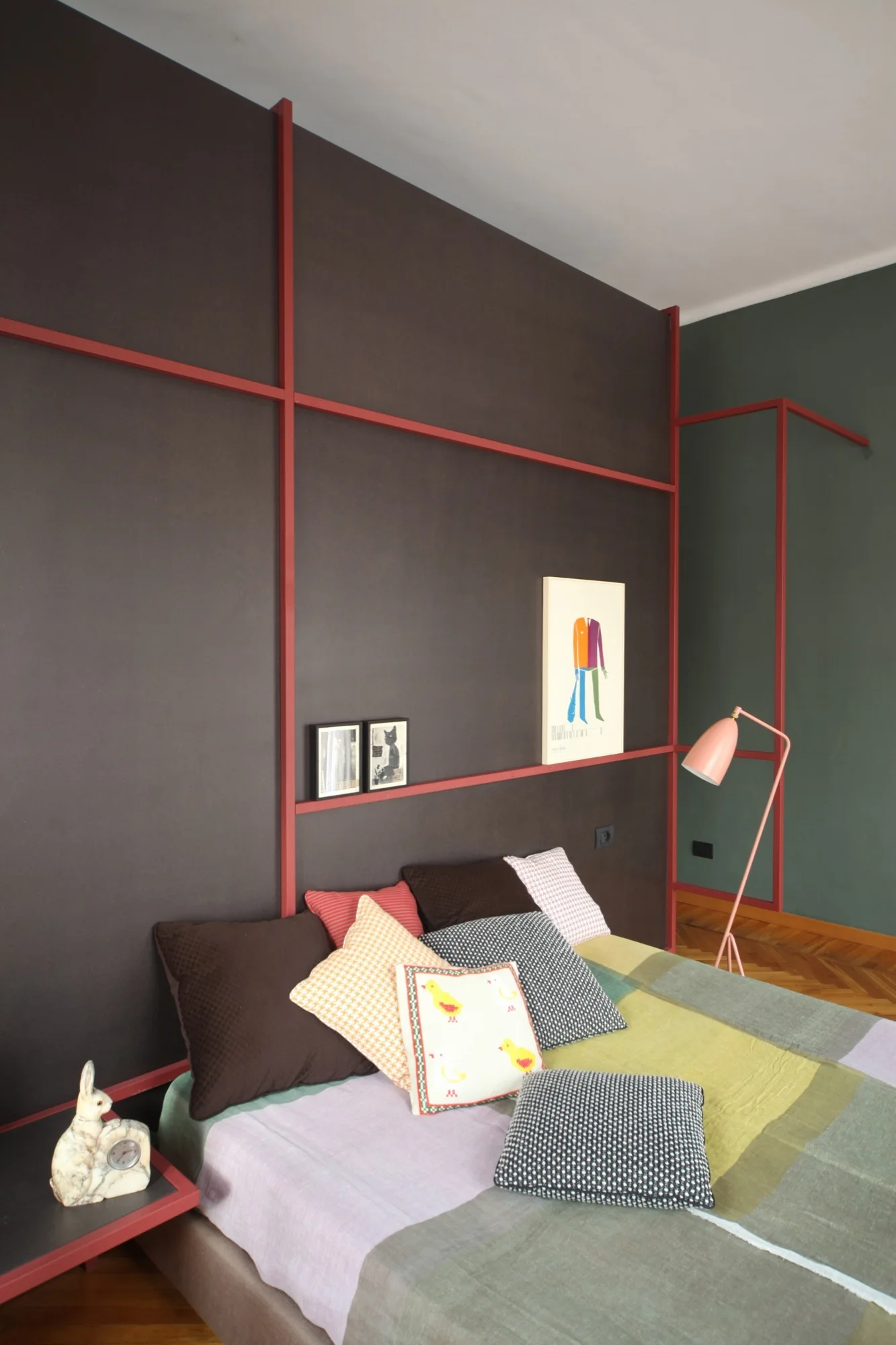 A bedroom features a bed with colorful patterned pillows against a dark brown wall accented with geometric red trim.