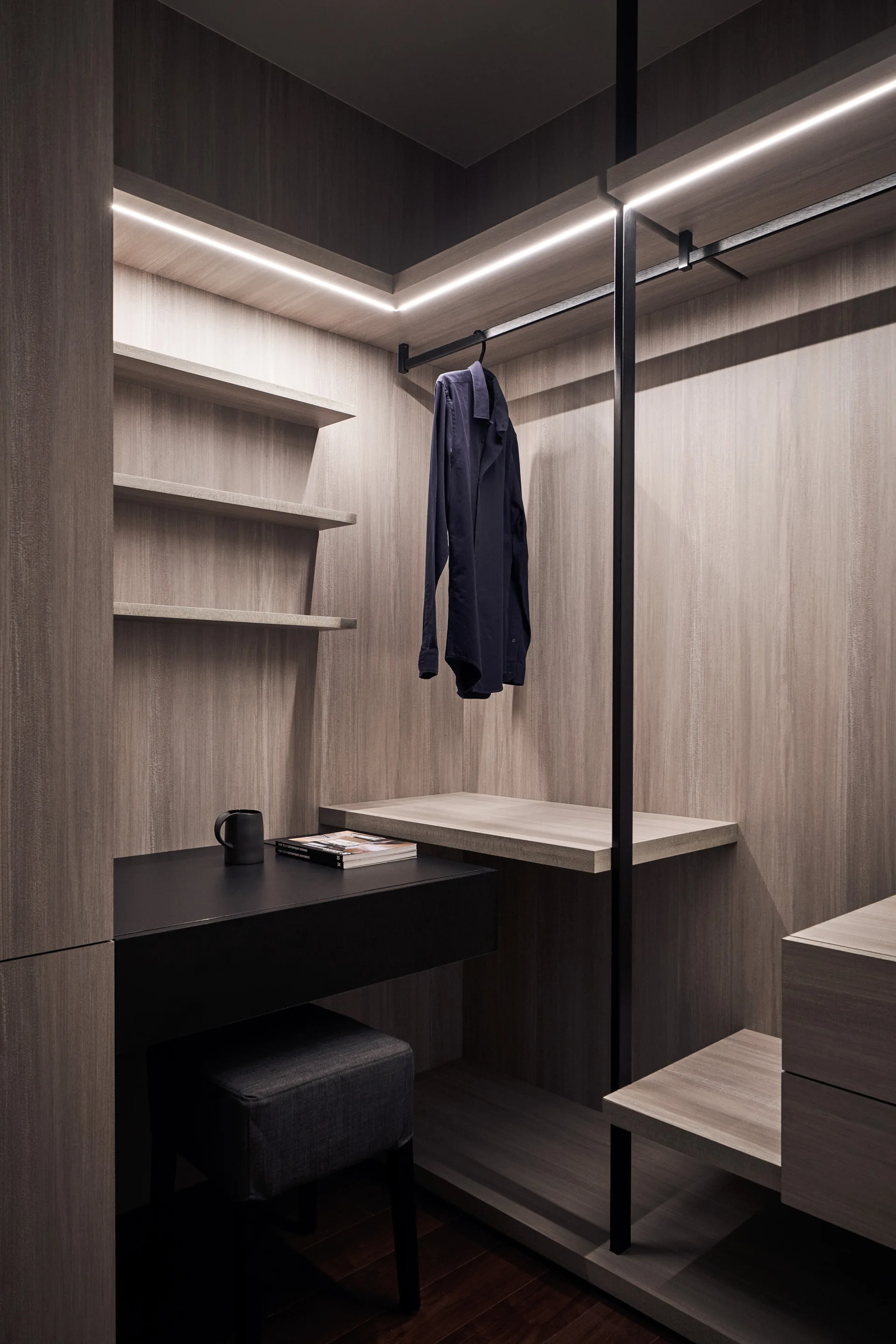 A modern walk-in closet featuring light wood paneling, built-in shelves, an integrated black desk with a small grey stool, and glowing LED shelf lighting.