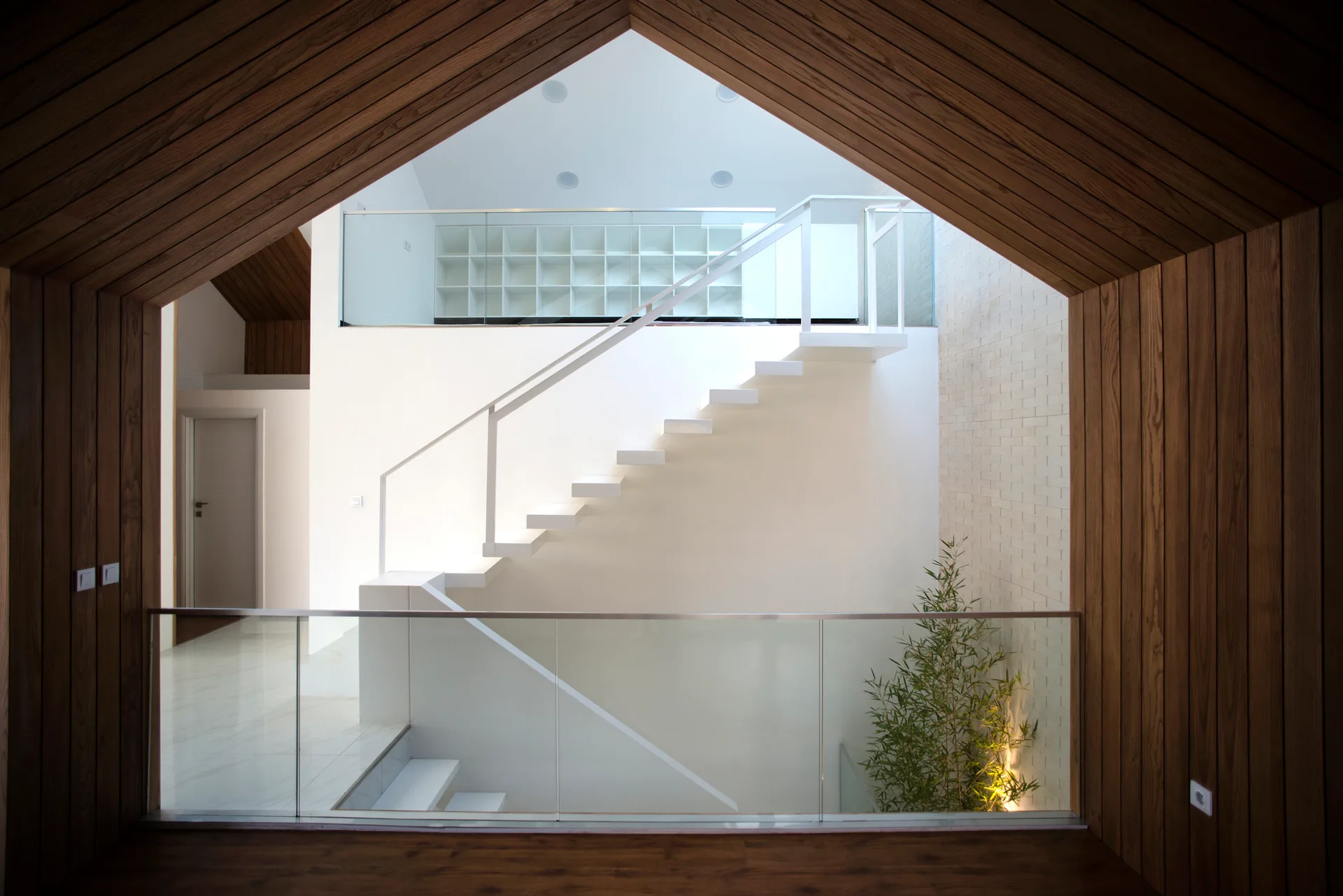 A white floating staircase with a glass railing is framed by a pitched, wood-paneled opening in a modern home.
