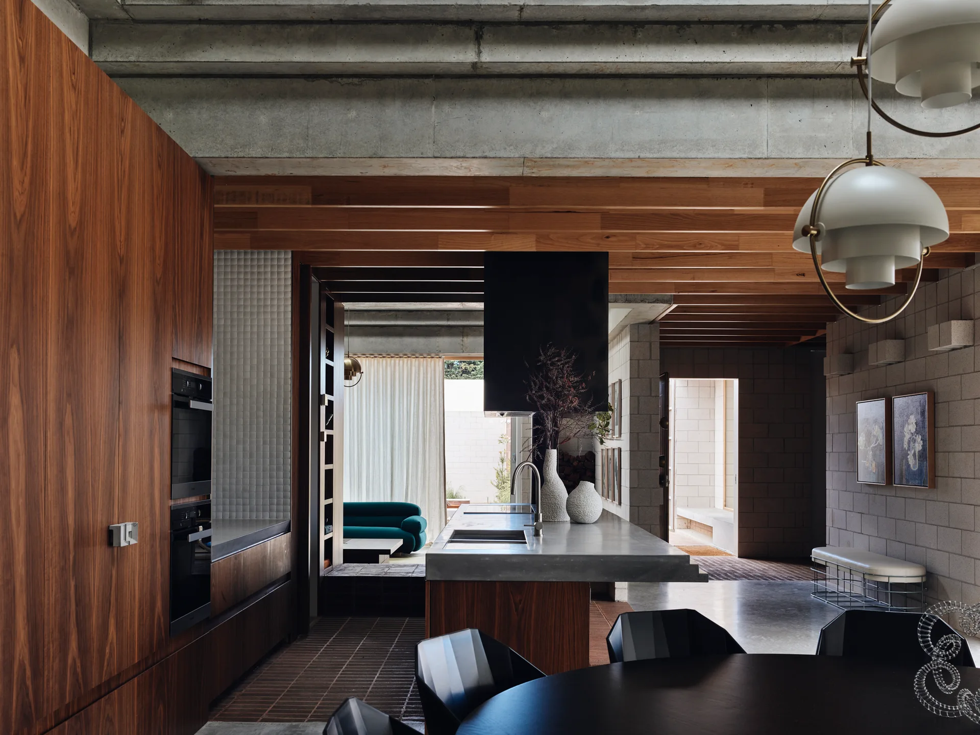 A modern kitchen features a central concrete island, warm wood-toned cabinetry, exposed concrete ceiling beams, and a view into a living area.