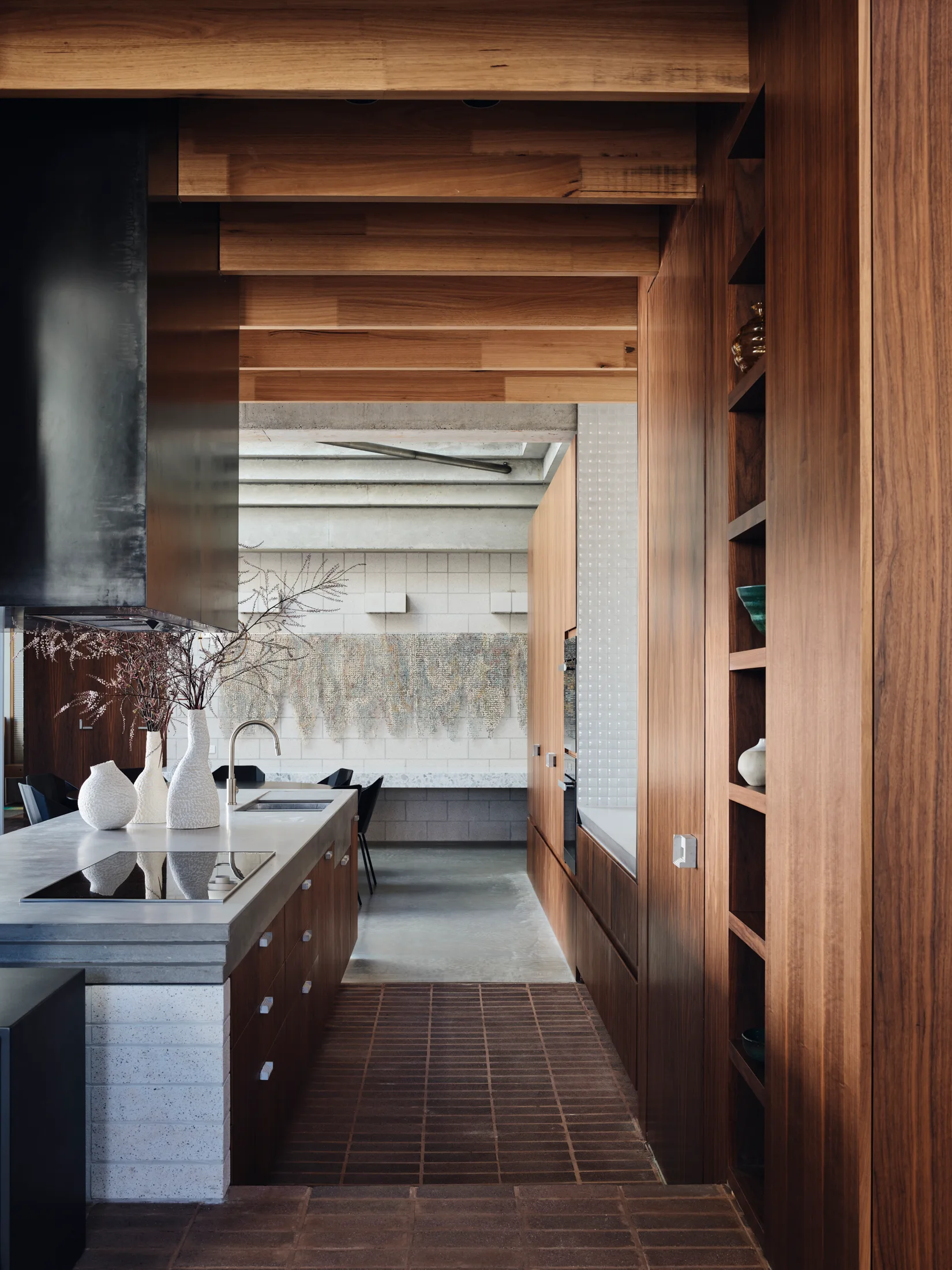 A modern kitchen features a concrete-topped island with textured vases, tall wood cabinets, exposed ceiling beams, and red brick flooring.