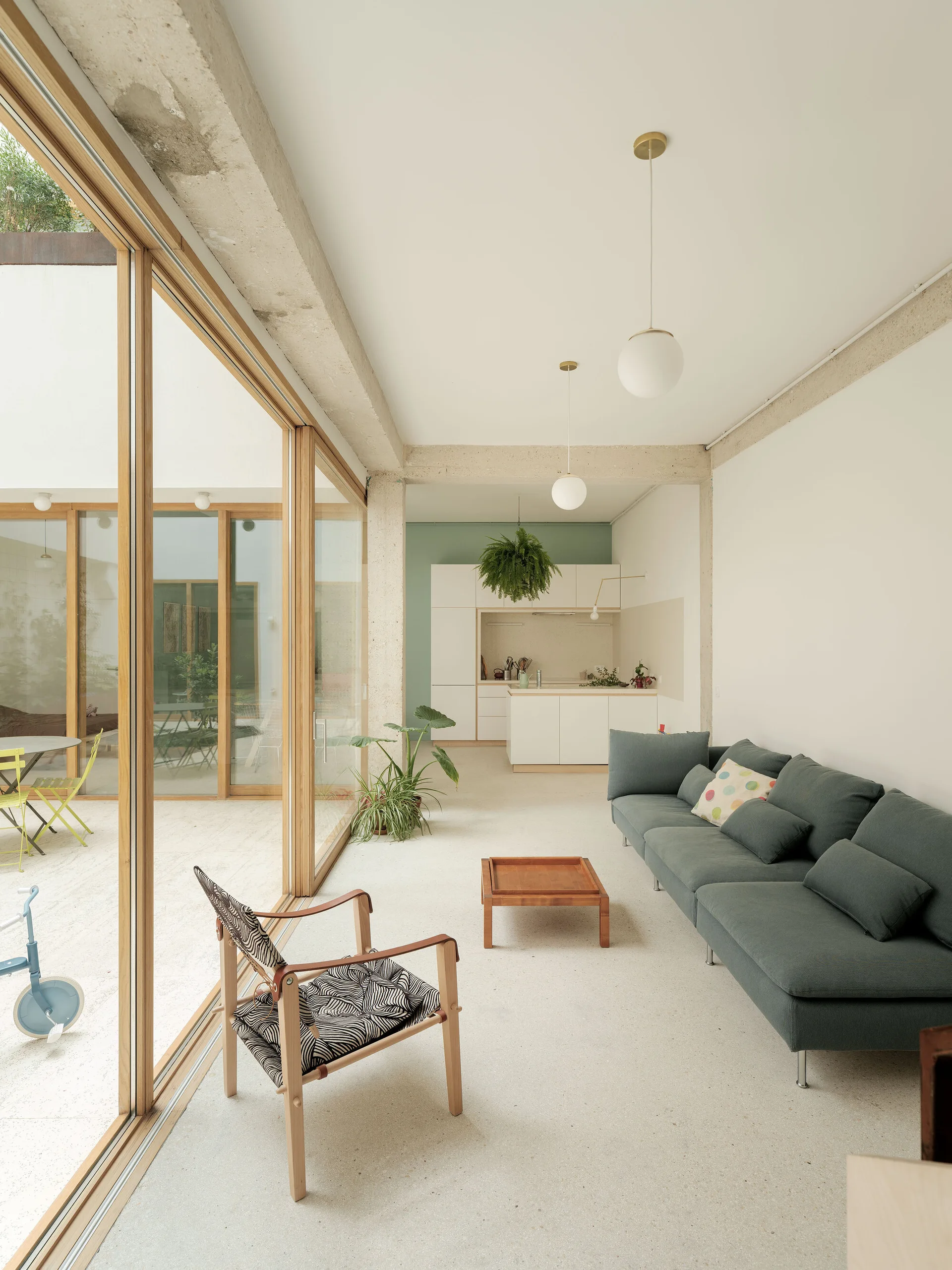 A minimalist living room with a grey sofa, wood-framed armchair, and large sliding glass doors opening onto an outdoor patio.