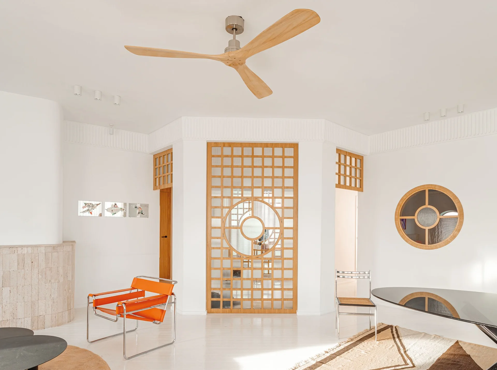 A bright white room featuring a wooden lattice room divider, a circular window, an orange strap armchair, and a light wood ceiling fan.