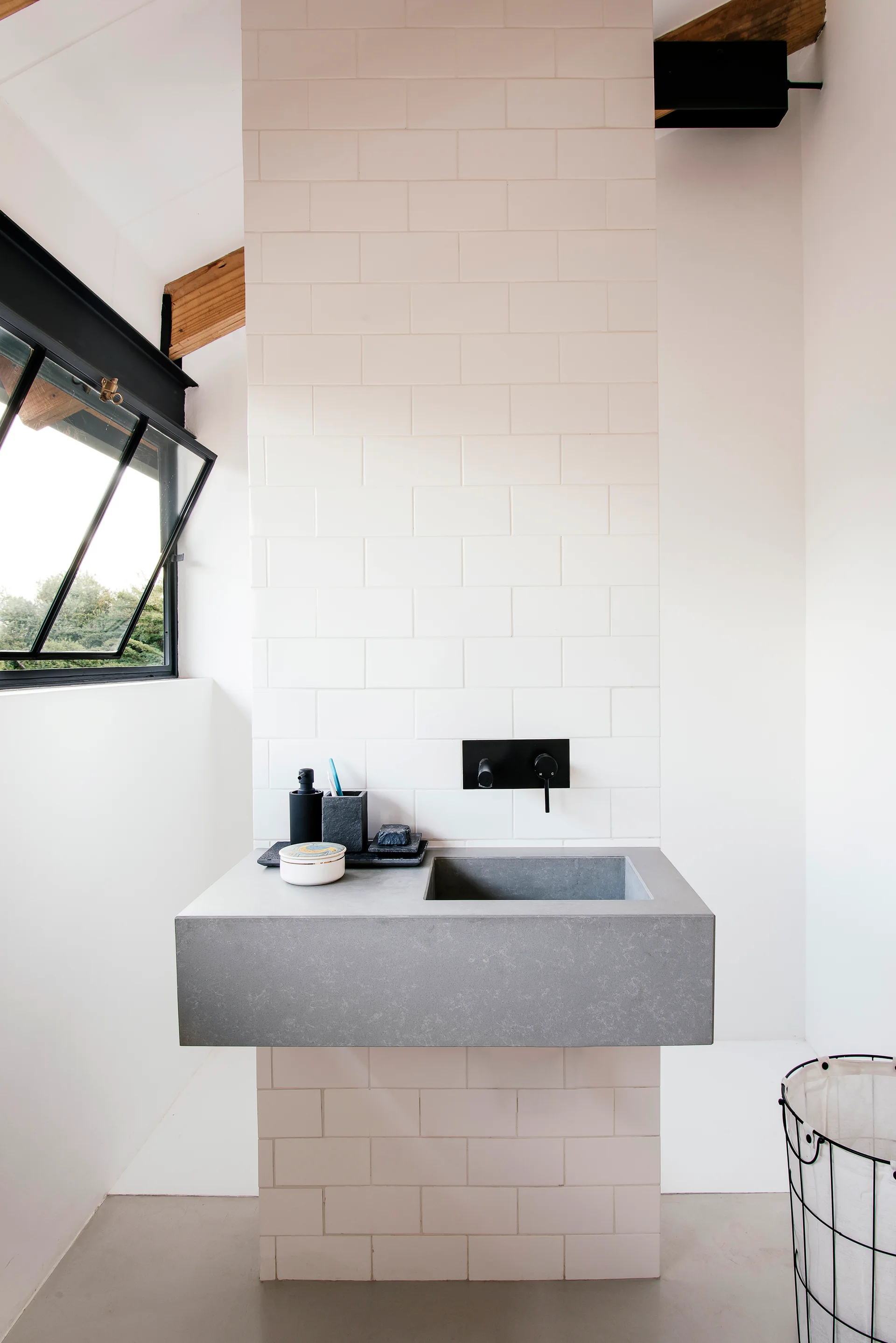 A grey concrete sink floats against a white subway tile wall with a matte black faucet and an open angled window.
