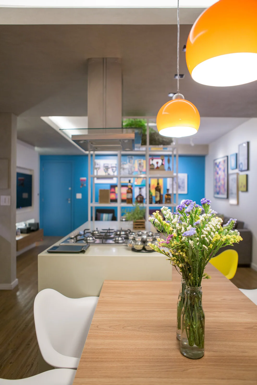 A dining table with flowers stands before a kitchen island with orange pendant lights and a vibrant blue accent wall in the background.