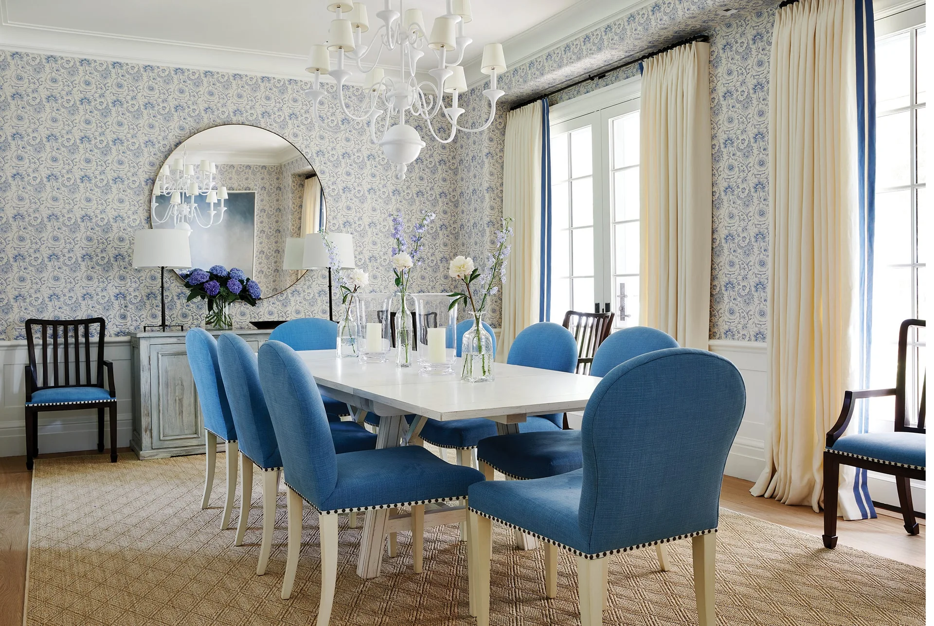 A traditional dining room features blue upholstered chairs around a white wooden table, set against blue floral wallpaper and white wainscoting.