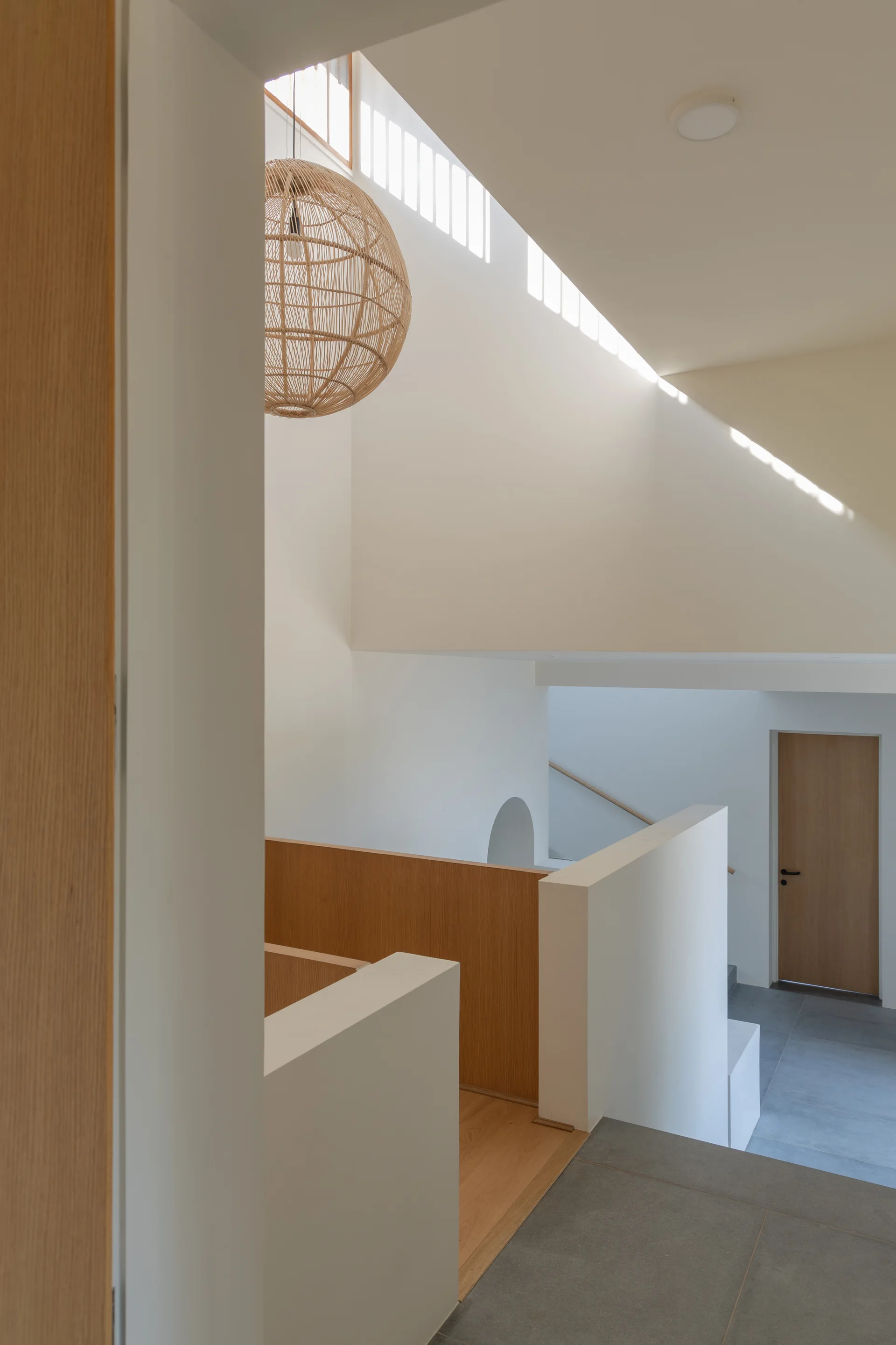 A modern minimalist staircase featuring smooth white walls, solid wood railings, grey tiled floors, and a large round woven pendant light.