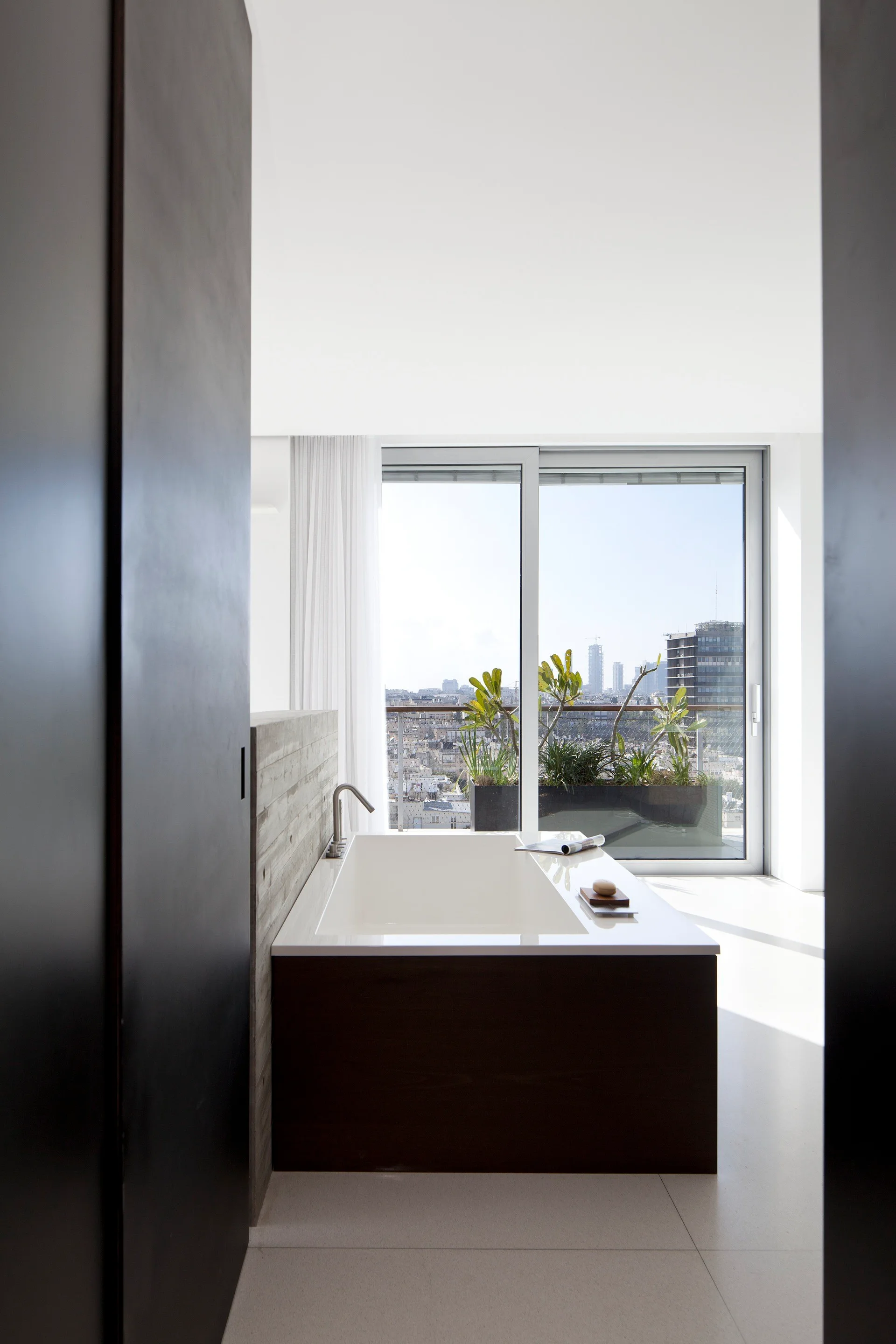 A modern bathroom with a white rectangular bathtub, a textured concrete feature wall, and large glass sliding doors looking out to a city terrace.