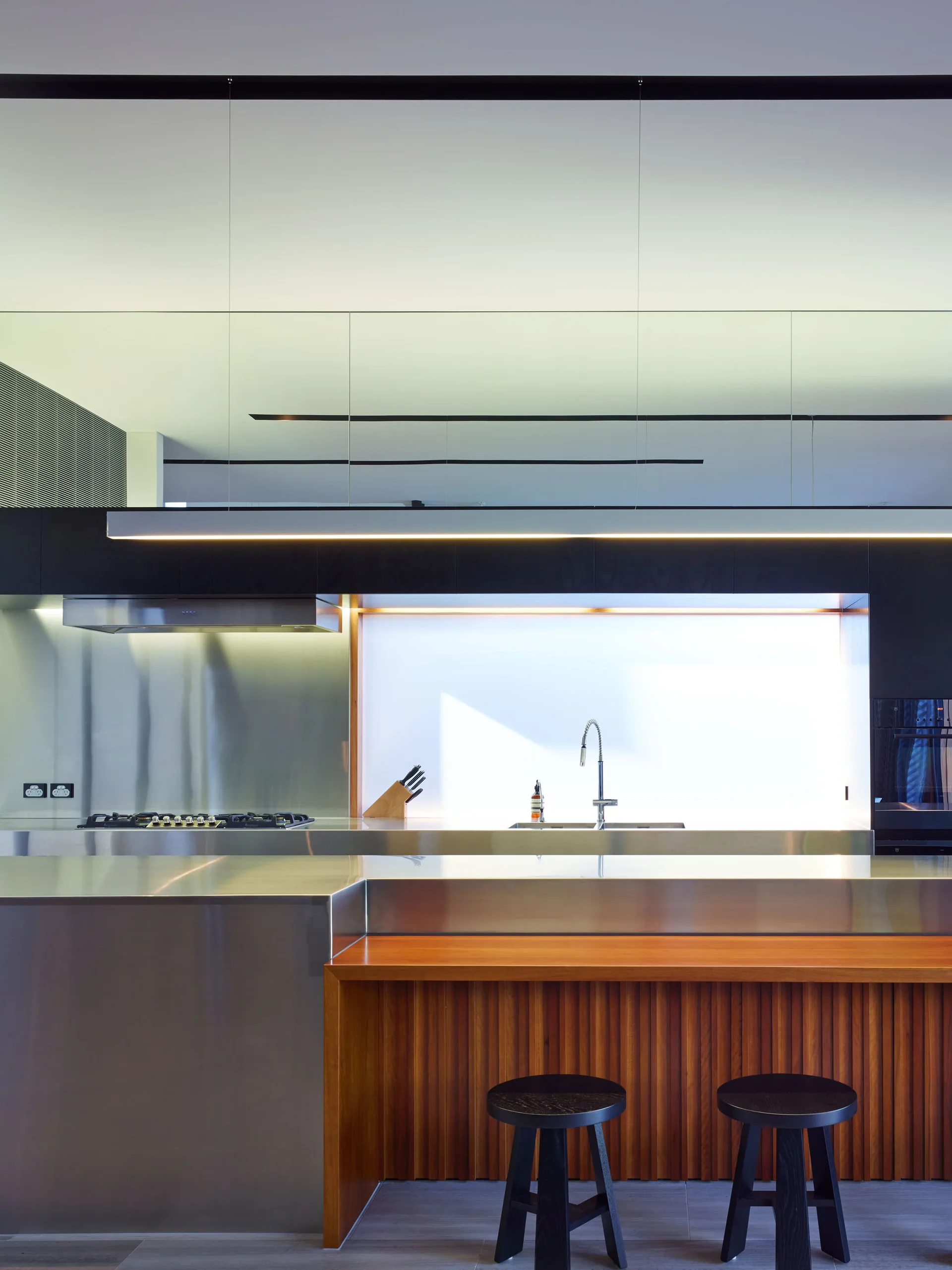 A modern kitchen featuring a stainless steel island with a slatted wooden base, black bar stools, and a glowing backlit backsplash.