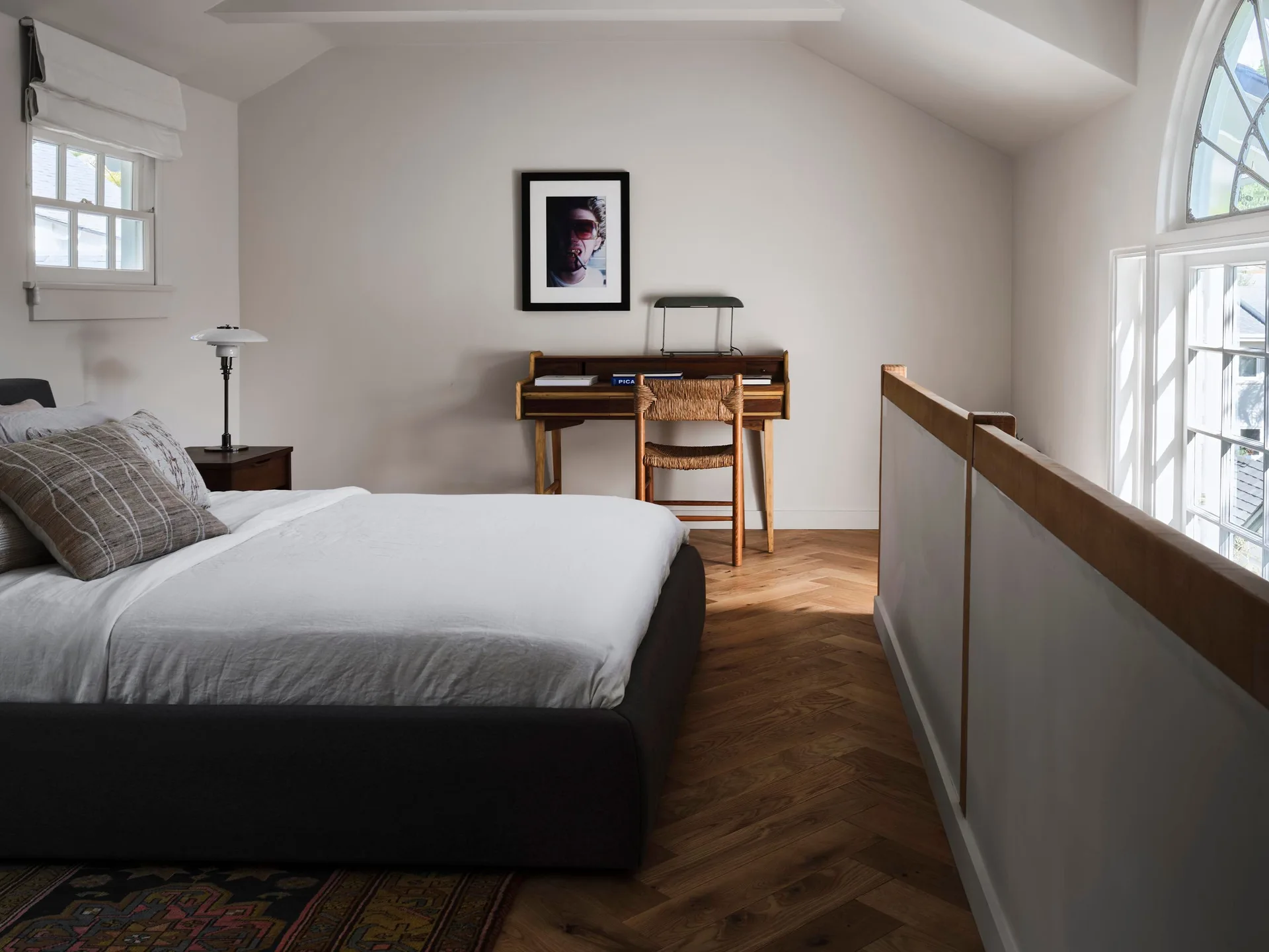 A bedroom features a dark upholstered bed, a mid-century wooden desk, and herringbone-patterned wood floors beside a half-wall railing.