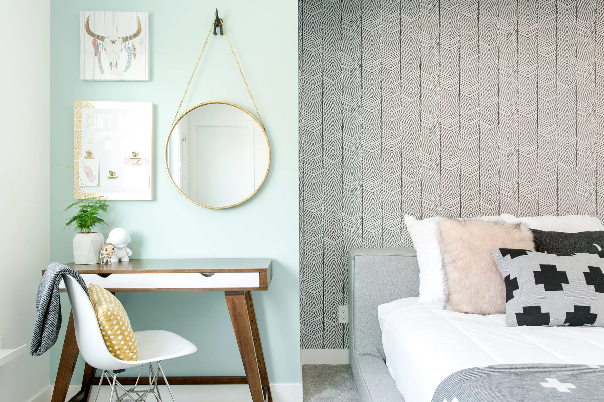 A modern bedroom features a wood and white writing desk against a mint green wall next to a bed with herringbone patterned wallpaper.