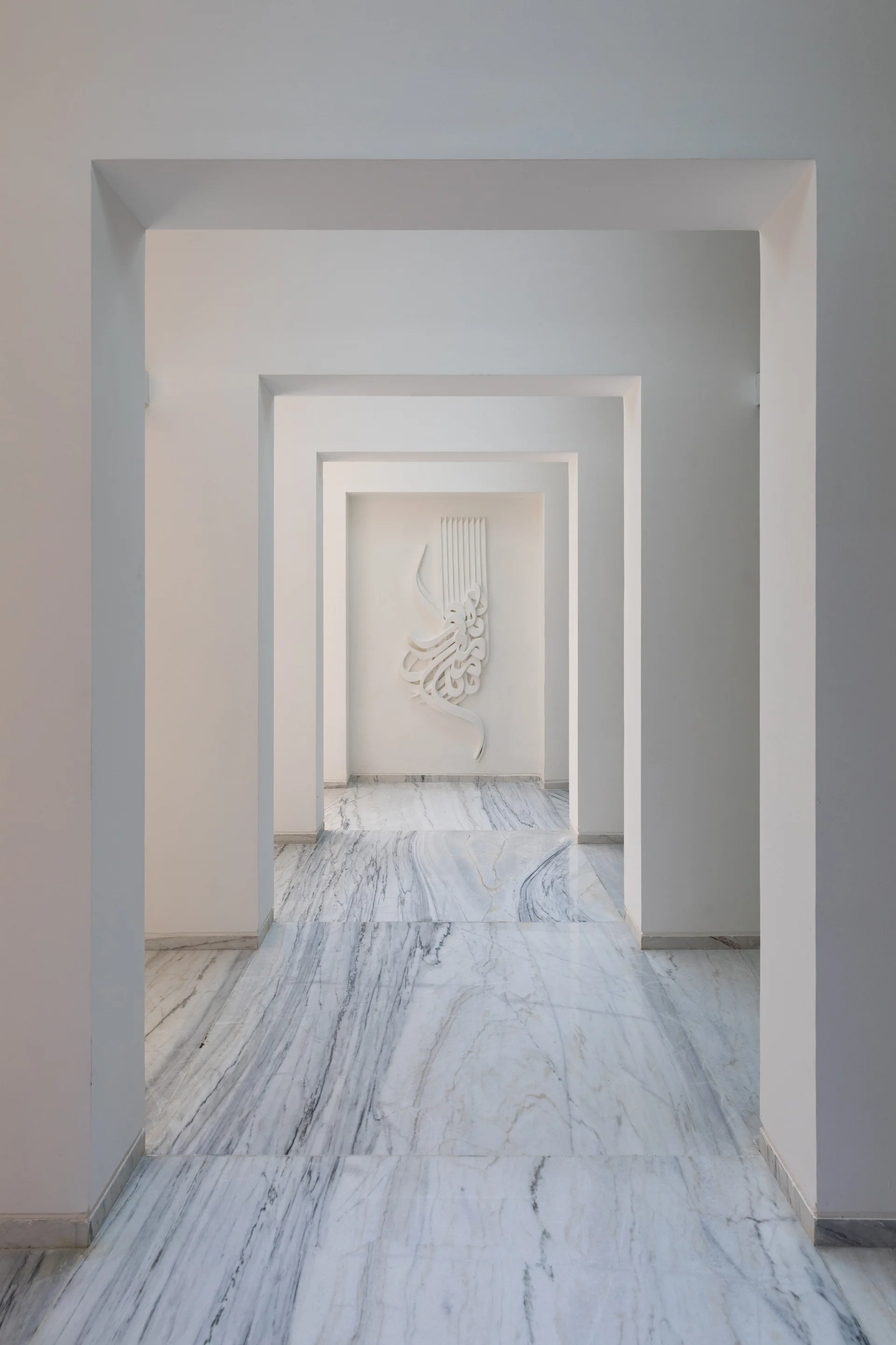 A long white hallway with repeating square archways leads to a sculptural calligraphy art piece on the back wall above polished marble floors.